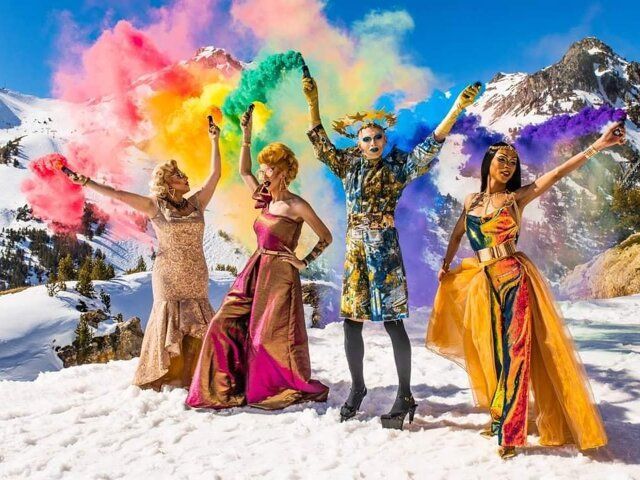Four people in colorful gowns holding smoke flares, snowy mountains background.