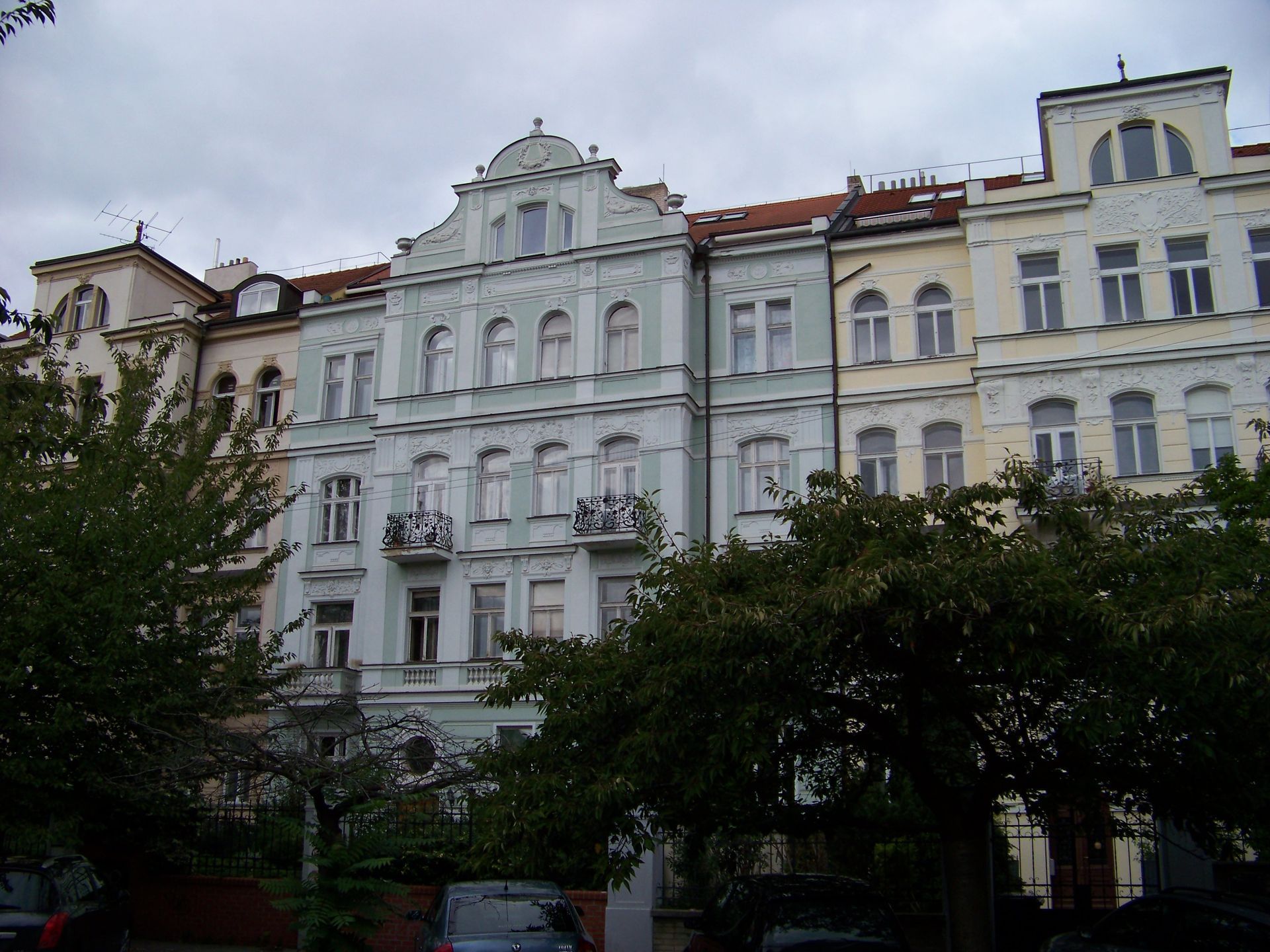 Row of colorful apartment buildings with architectural details, trees in front, overcast sky. Vinohrady Prague