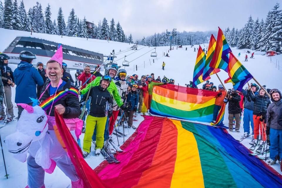 Group of skiers on snowy mountain holding rainbow flags and celebrating, some in costume. Whistler Pride