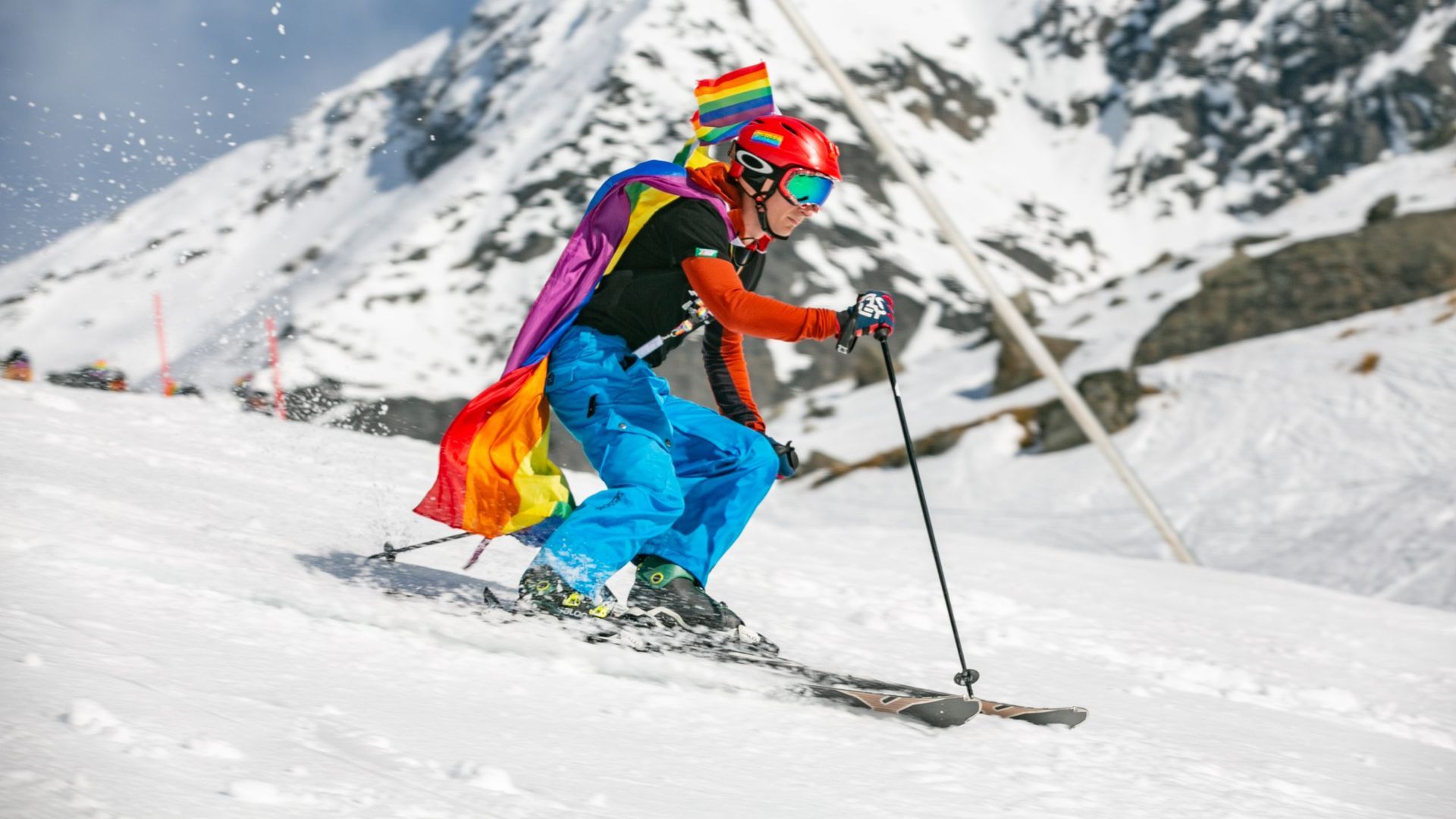 Skier on snowy slope, wearing rainbow cape and helmet. Mountains in background. Winter Pride New Zealand