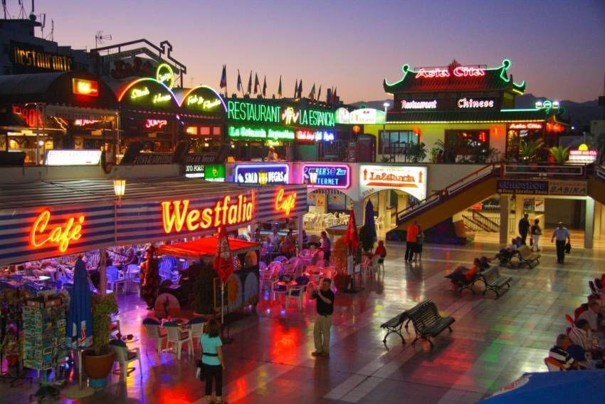Night scene of a plaza with restaurants and shops lit with neon signs. People sit and walk about. Yumbo Centre
