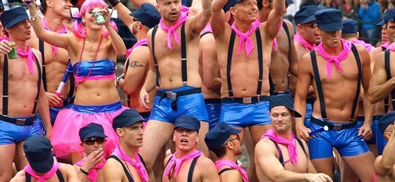 A group of people wearing blue shorts, black suspenders, and pink neck scarves, posing in an outdoor parade setting.