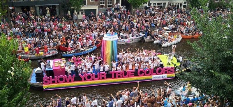A canal filled with boats during a Pride parade, featuring a float with a rainbow flag and the text 