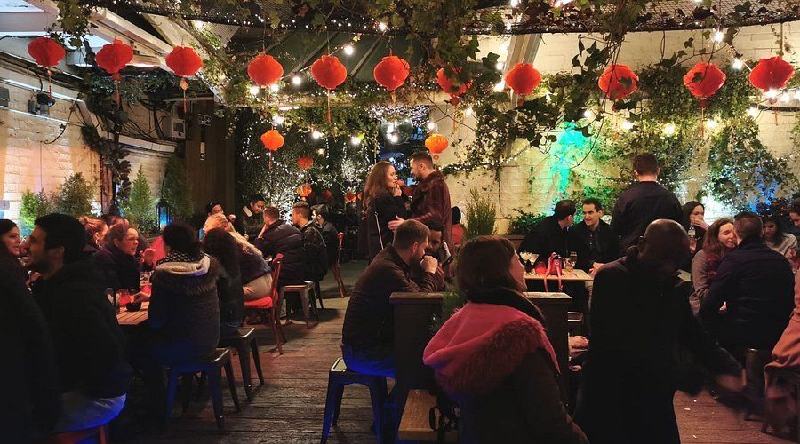 Outdoor gathering, people seated at tables, red lanterns, string lights, greenery overhead, festive atmosphere. The Yard