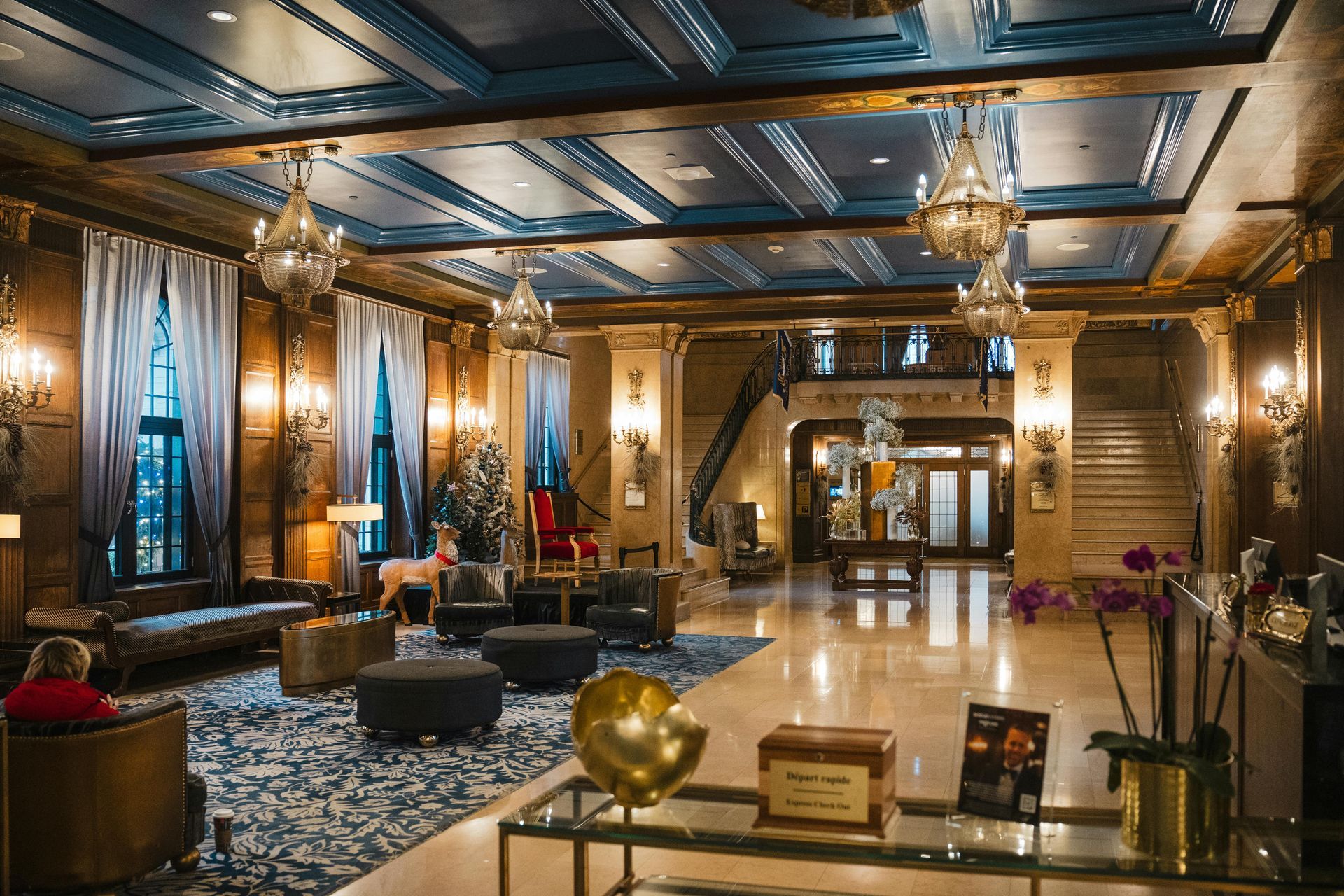 A luxurious hotel lobby with dark blue coffered ceilings, ornate chandeliers, plush seating, and marble floors.