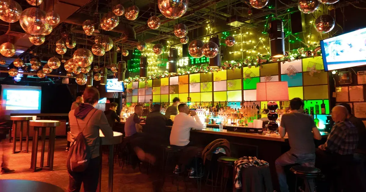 Bar interior with disco balls, patrons at counter, and colorful backdrop.