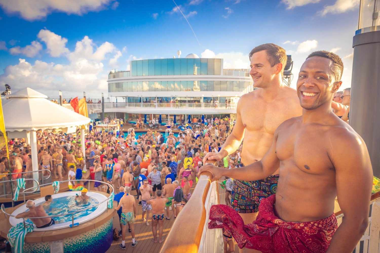 People on a cruise ship deck partying by a pool. Some are shirtless and smiling. Blue sky in background. Gay Cruise
