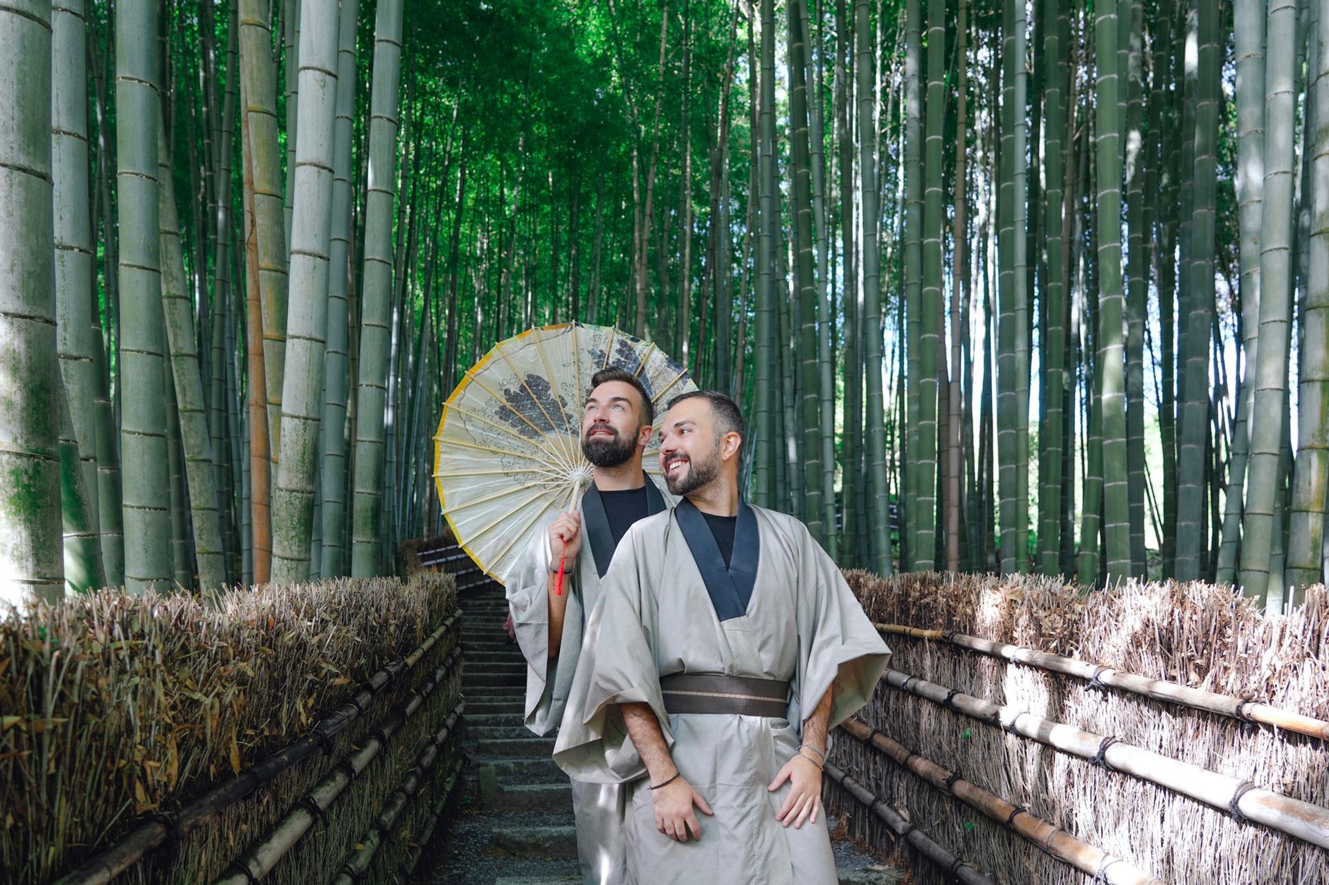 Two people in traditional Japanese attire stand in bamboo forest, one holding a parasol.