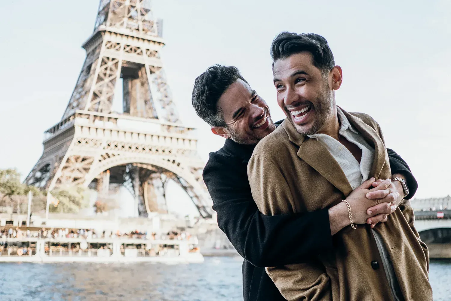 Two men laughing, hugging in front of the Eiffel Tower in Paris.