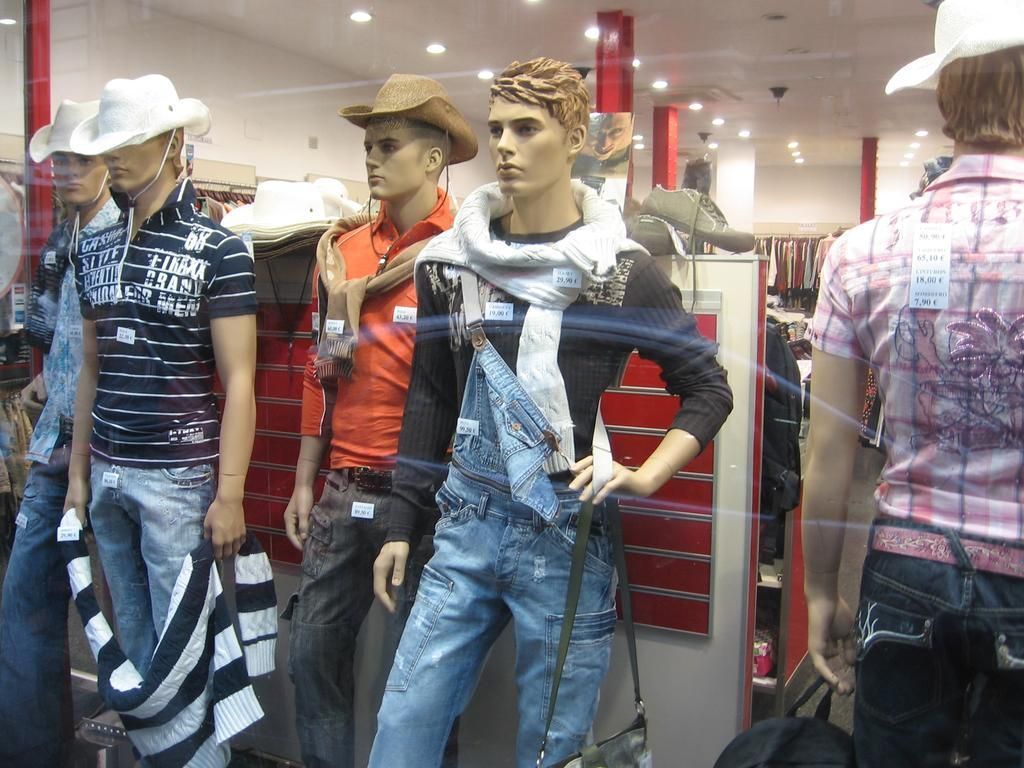 Mannequins in a store window display men's clothing: jeans, shirts, hats.