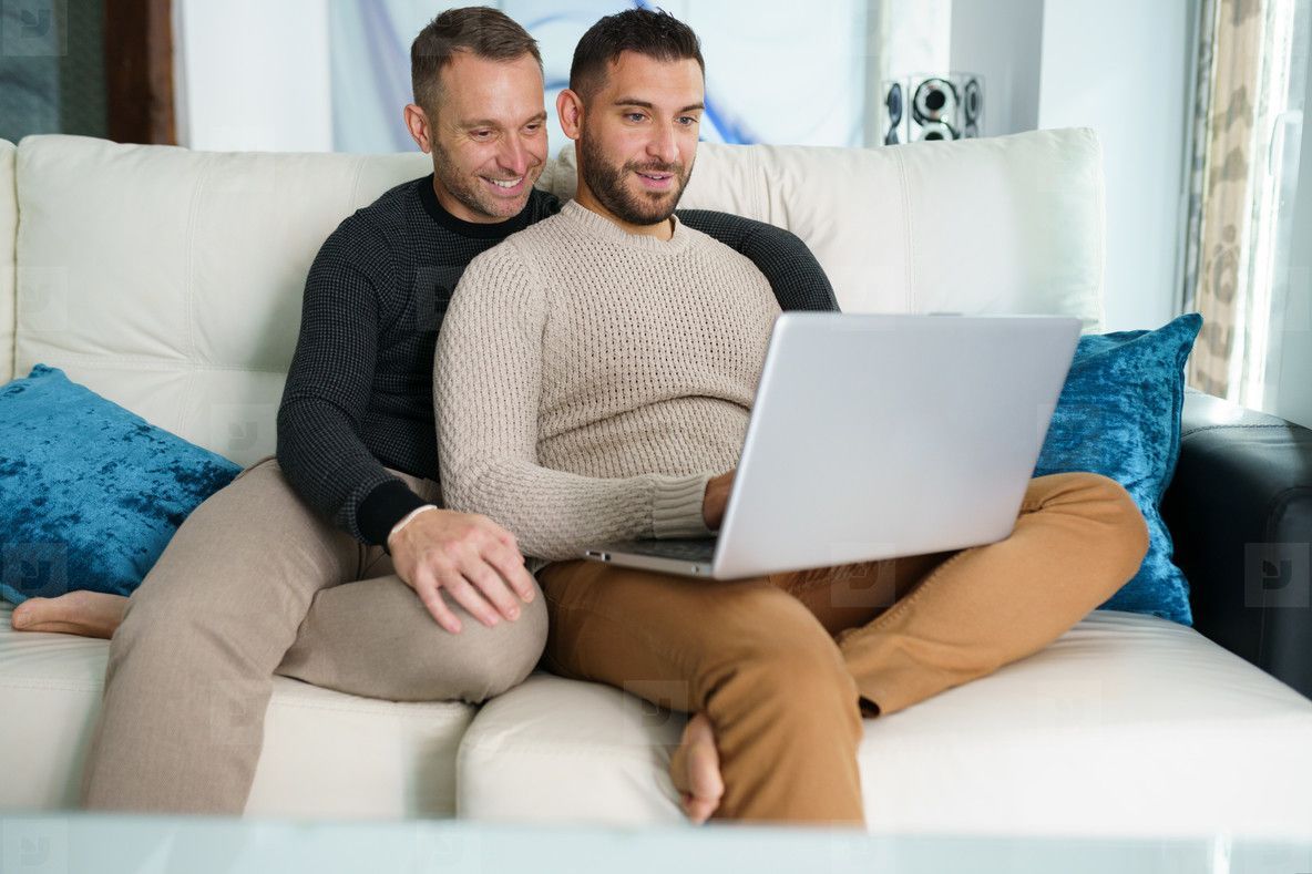 Two men sitting close together on a couch, looking at a laptop.