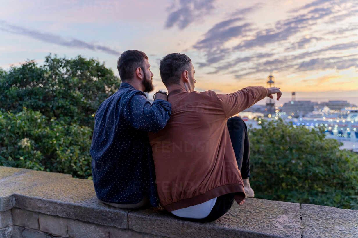 Two people sit on a wall, looking at a city sunset. One points, the other has an arm over their shoulder.