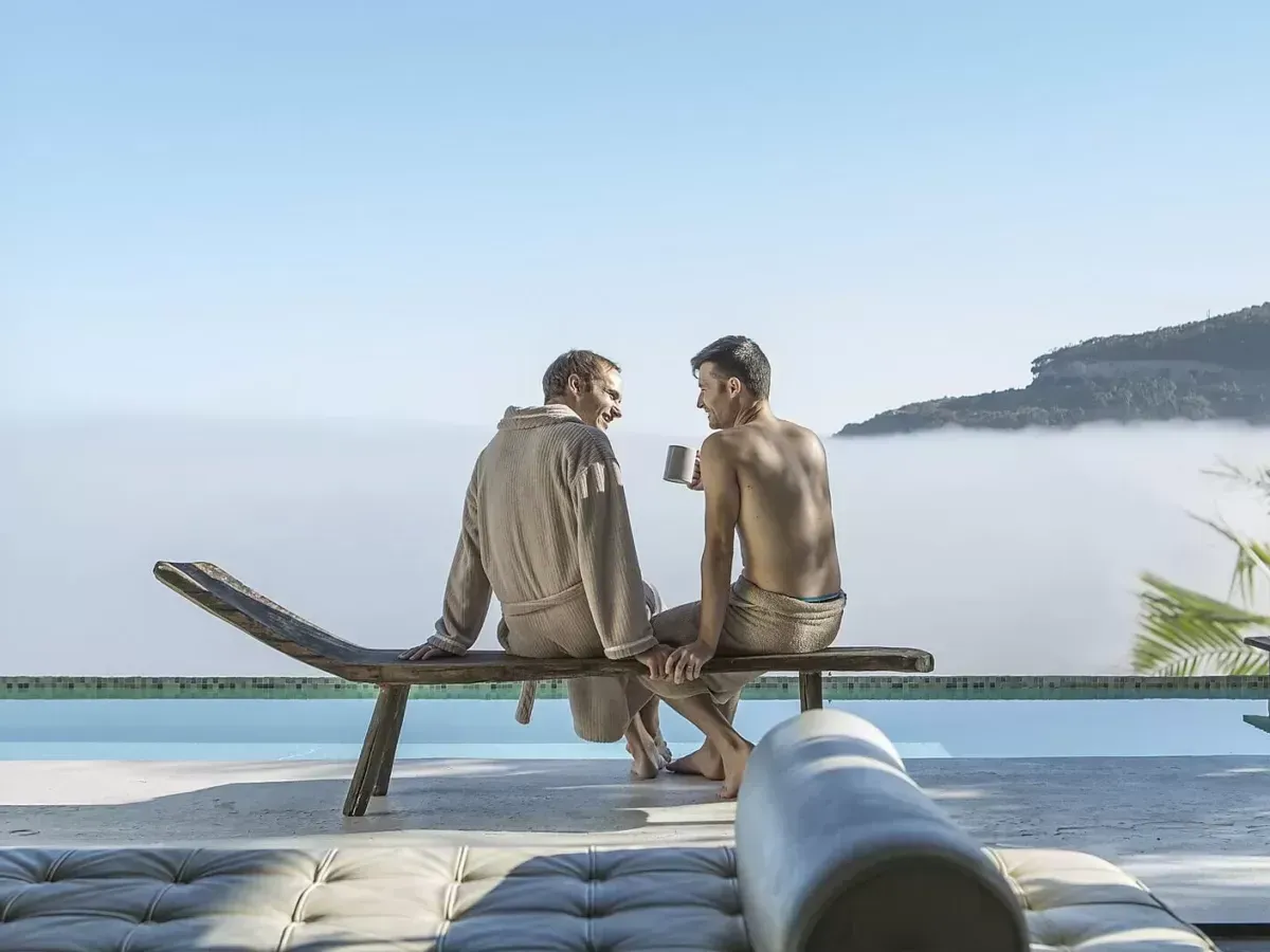 Two people sit on a wooden chaise lounge overlooking a misty landscape and swimming pool. One wears a robe.