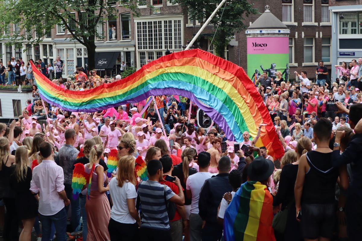 A large rainbow flag arches over a crowd gathered in an Amsterdam street for a Pride celebration.