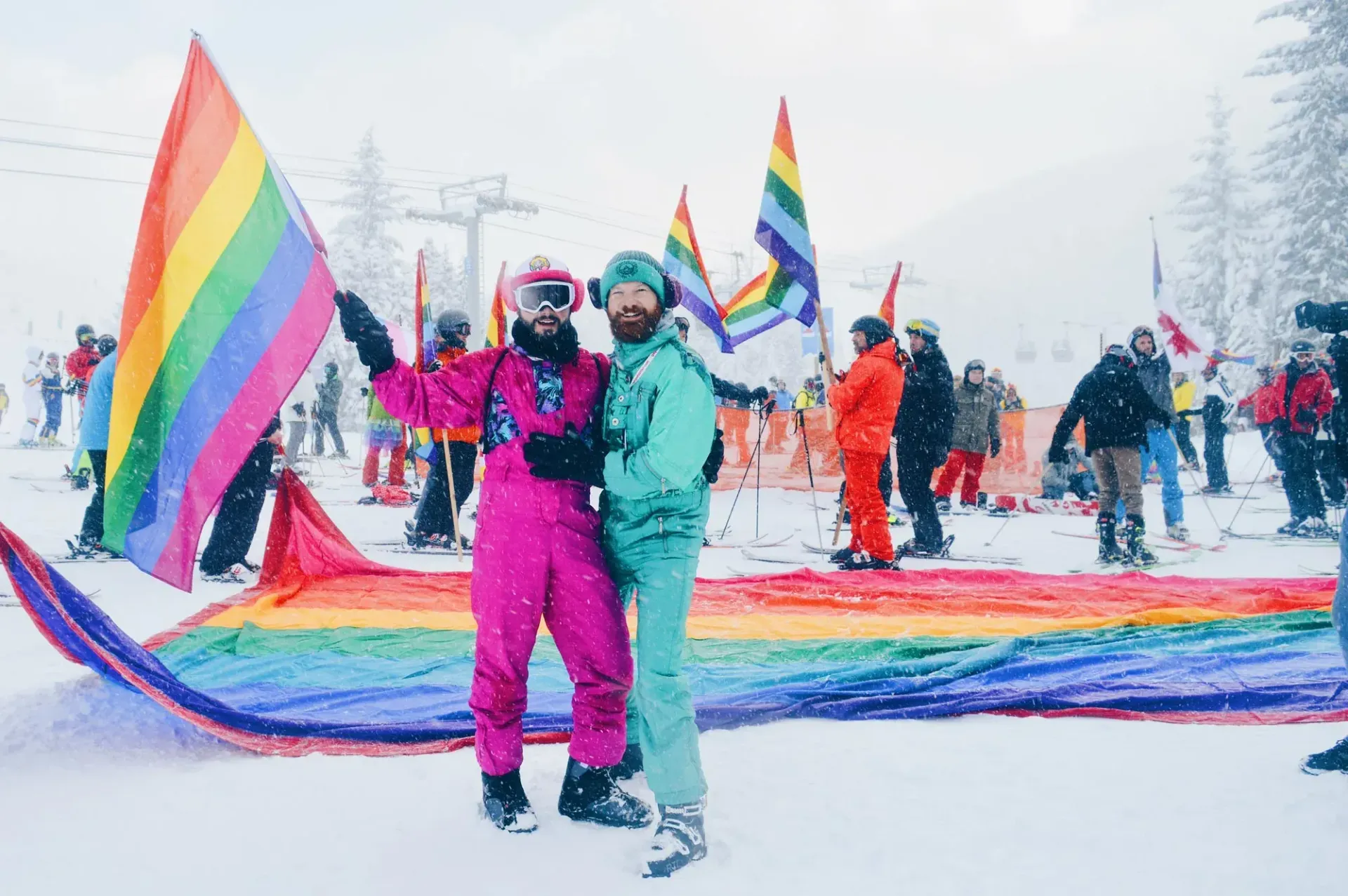 Two people in ski suits pose with LGBTQ+ flags and a rainbow in the snow.