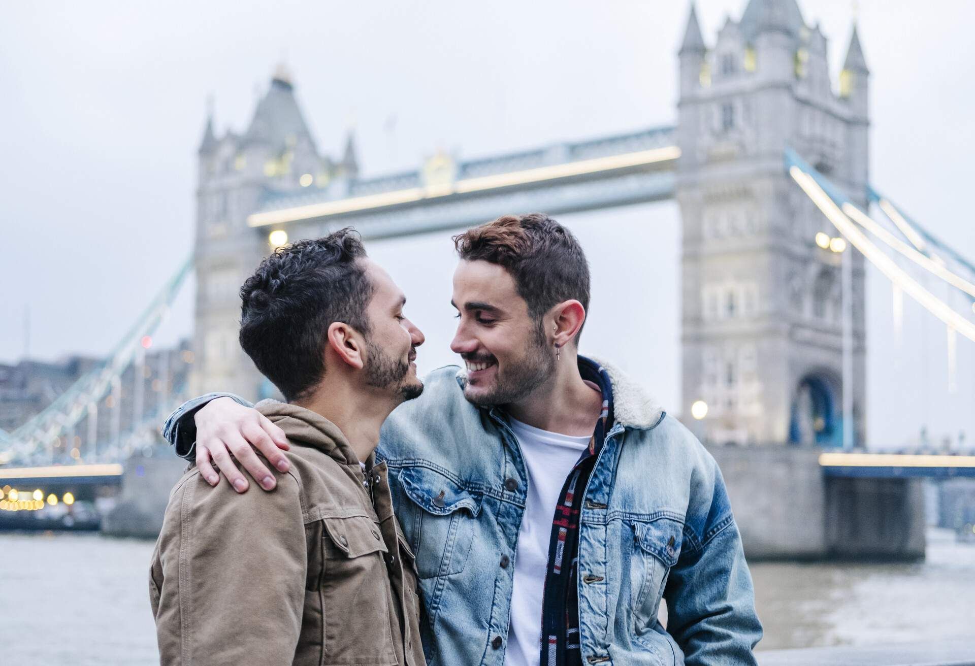 Two men embrace in front of Tower Bridge, London. One wears a denim jacket and smiles, looking at the other.