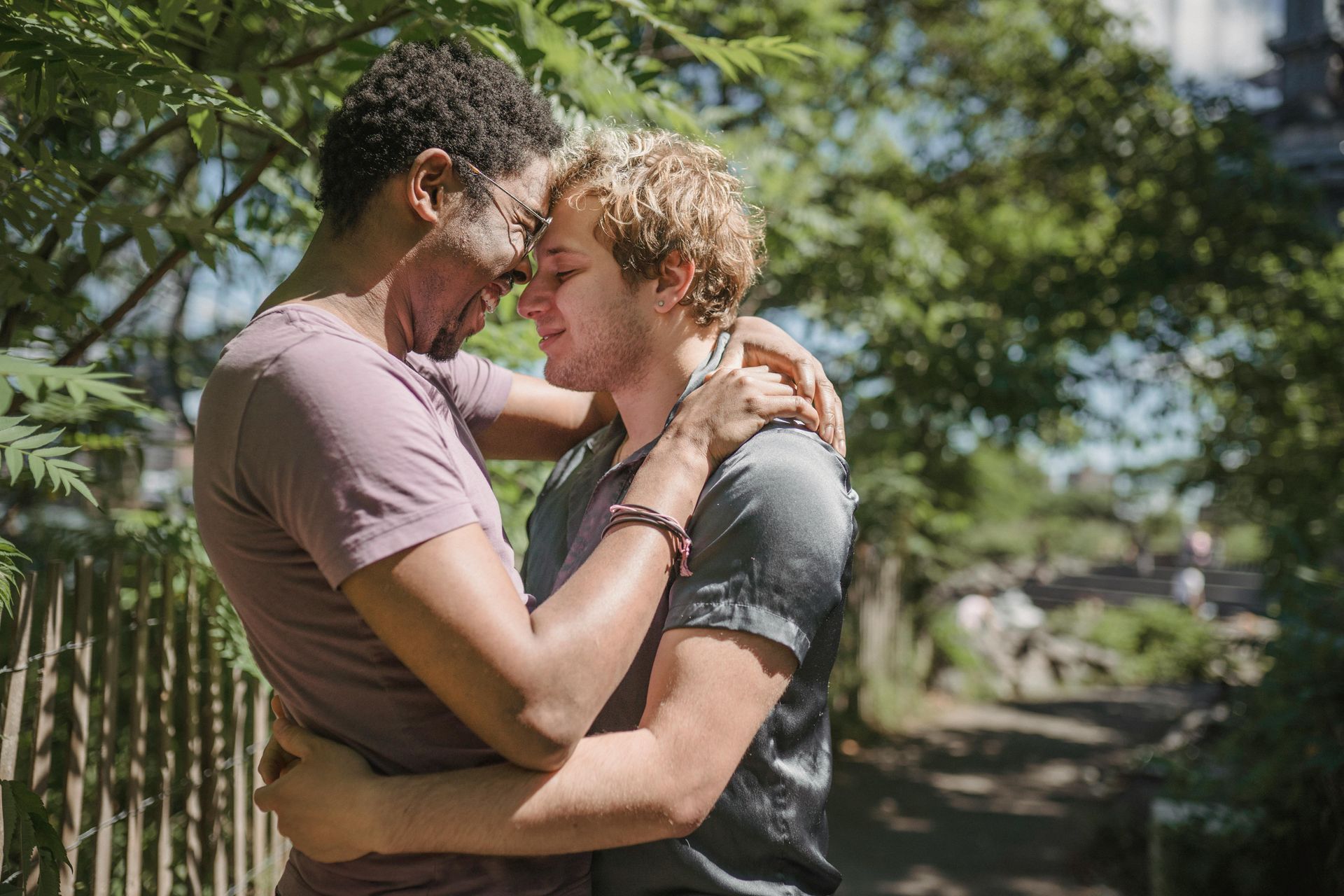 Two people embracing outdoors; one with dark skin, the other with light skin and blonde hair. Sunlight and greenery in background.