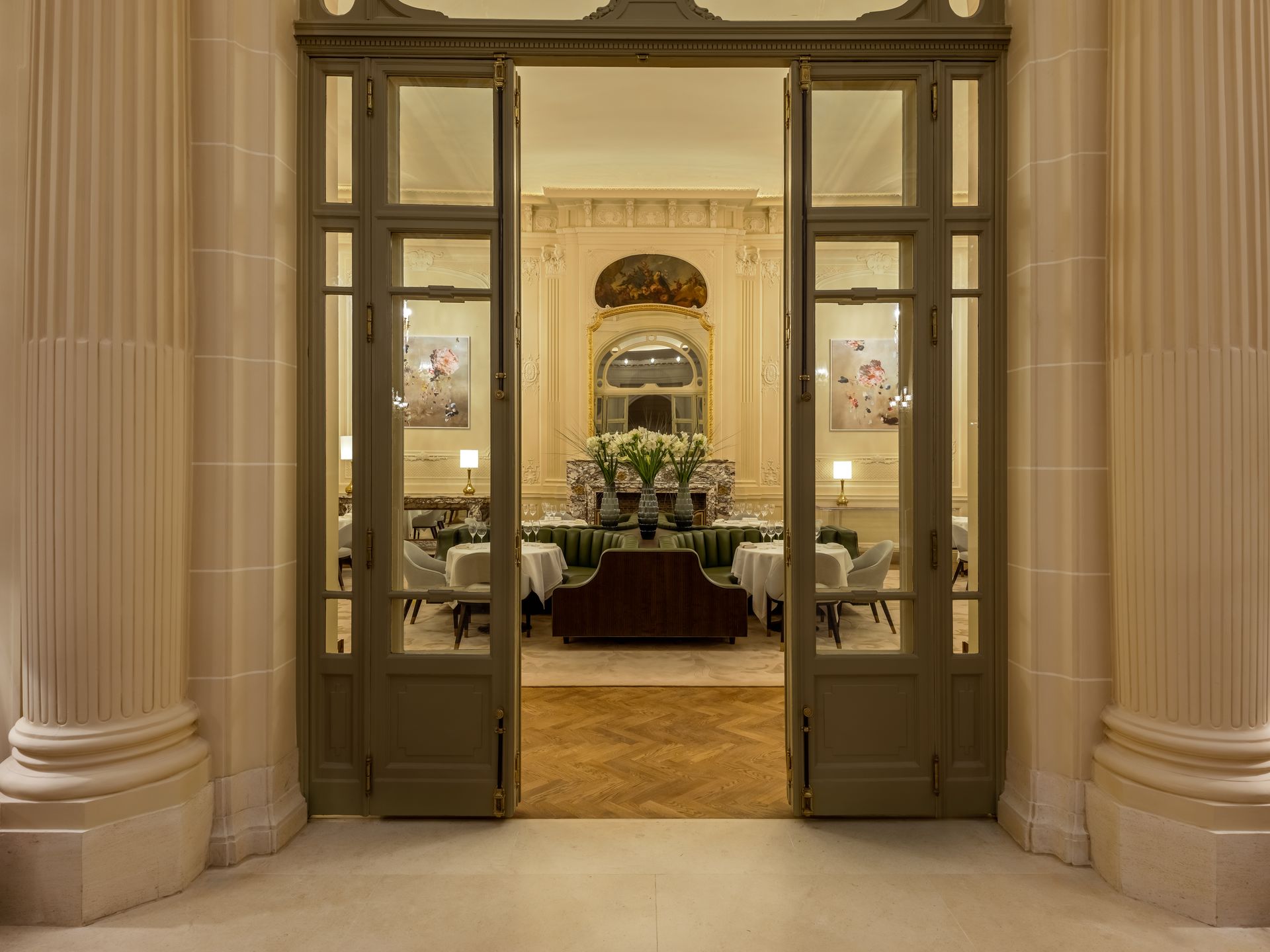 A wide doorway with open glass doors leads into a formal dining room with tables, chairs, and a central floral display.