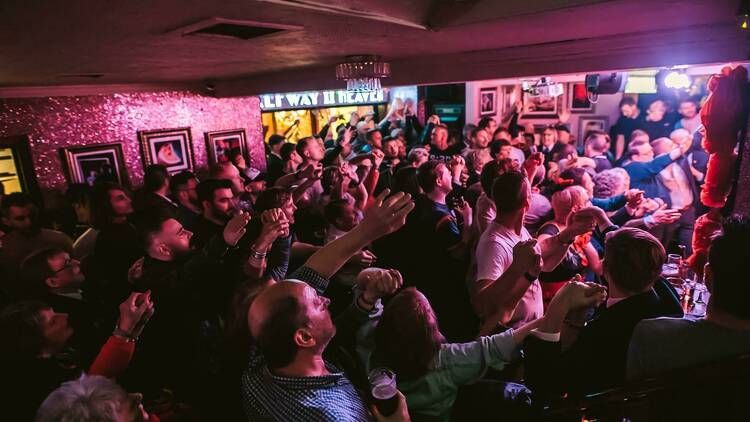Large crowd in a bar, arms raised, cheering. Dark, warm lighting; decorative walls, visible signs.