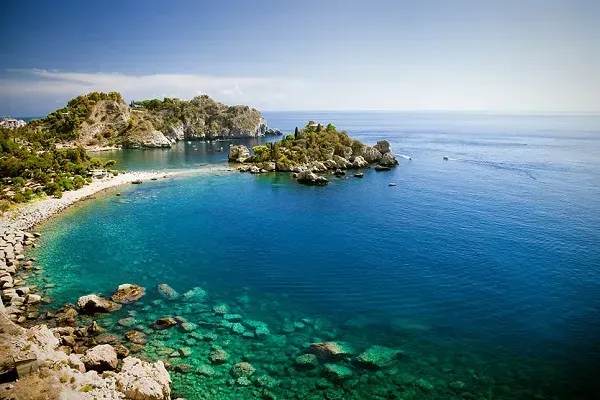 Azure waters surround a rocky coastline and small islands, trees and blue skies. Sicily