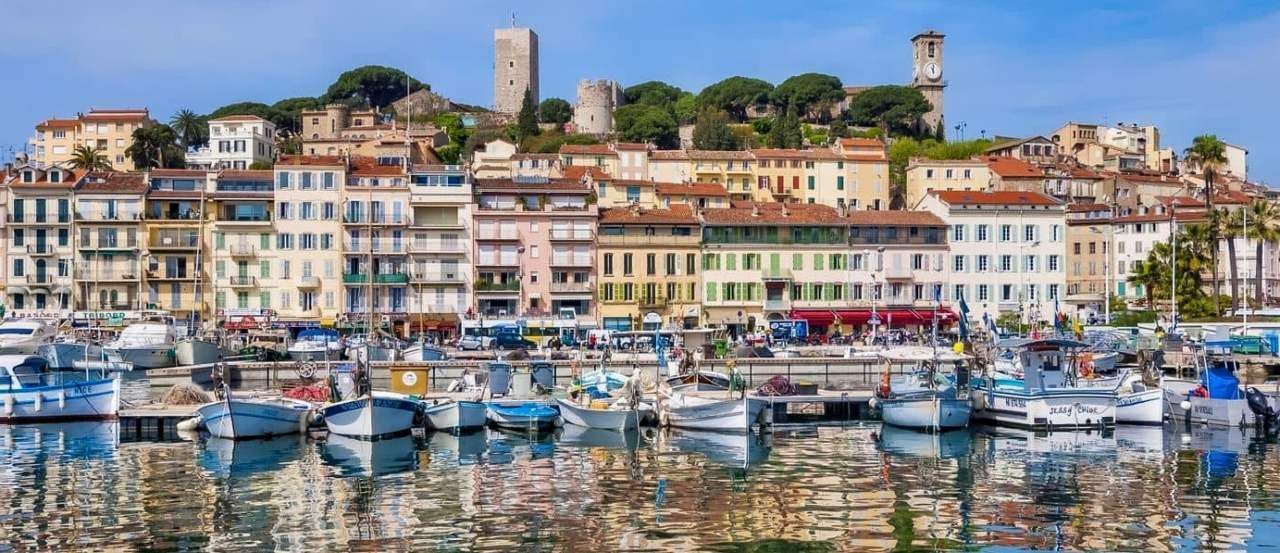 Boats in a harbor in front of a colorful seaside town. Buildings line the shore, topped by a tower. Cannes