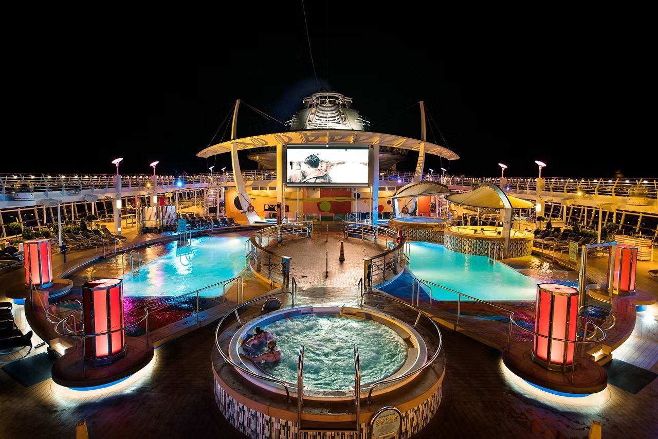 Nighttime view of a cruise ship deck with illuminated pools, hot tub, and a large screen under a dark sky.