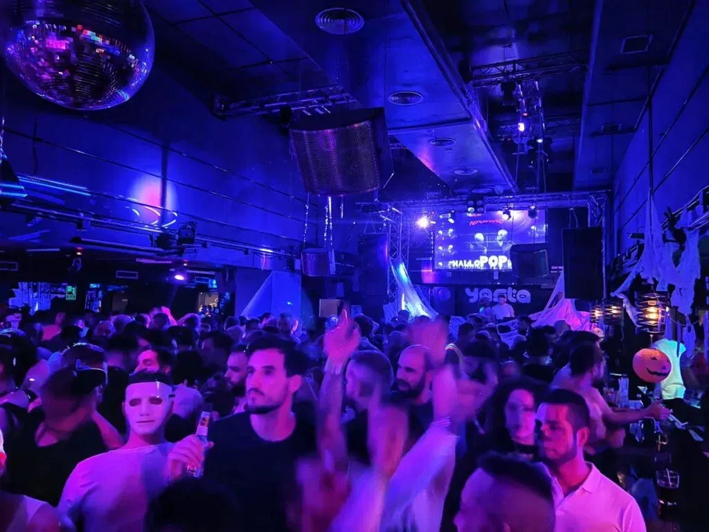Crowd in a dimly lit club with a DJ stage, disco ball, and blue lighting.
