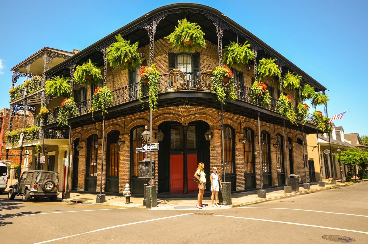 Corner building with wrought-iron balconies, hanging ferns, and red doors. Two women stand in front.