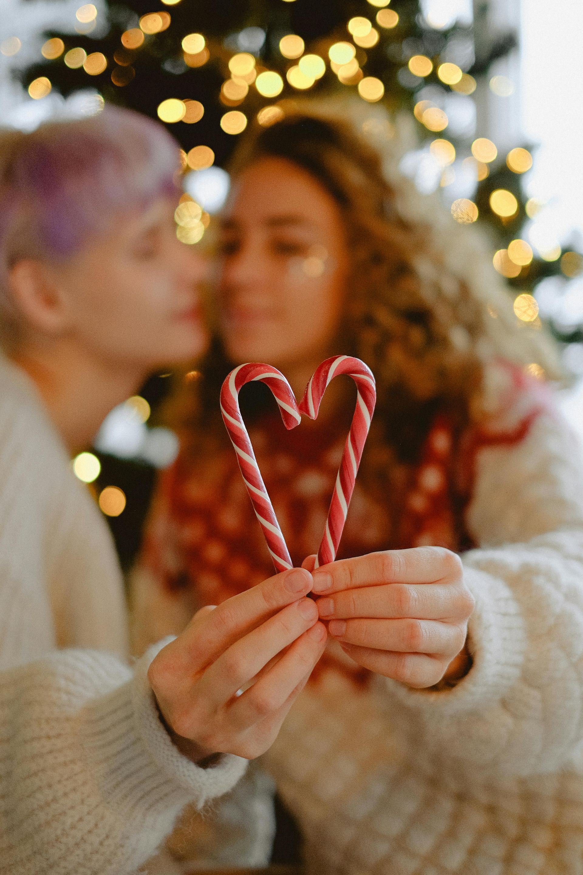Two people holding candy canes in a heart shape, with a blurred Christmas tree background.