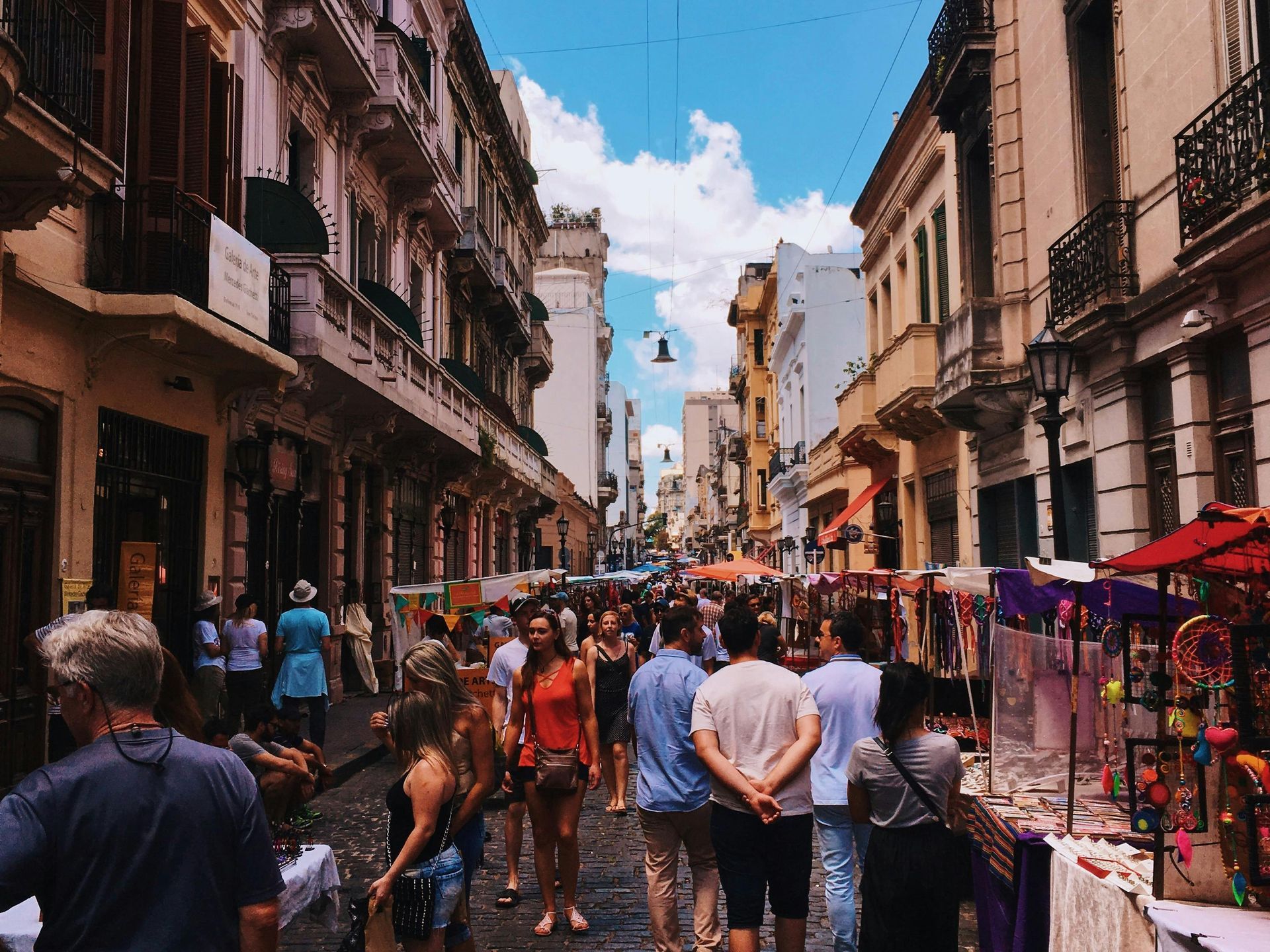 People stroll along a crowded street lined with shops and vendors under a blue sky. Buenos Aires