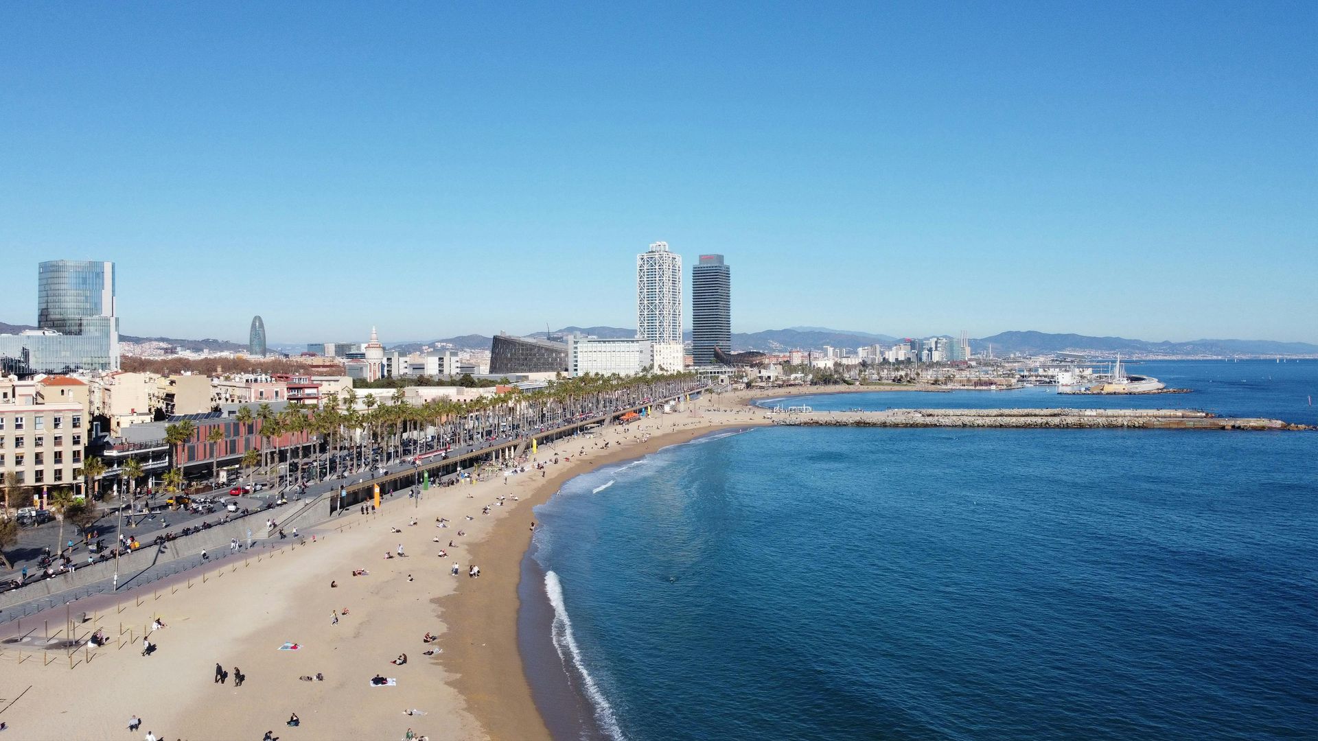 Beachfront in Barcelona with sand, blue sea, people, and city skyline under a clear, blue sky.