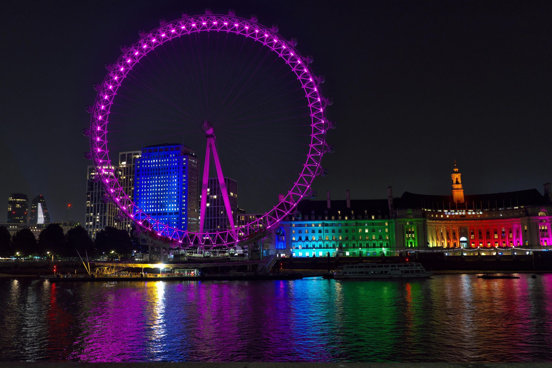 London Eye at night, lit in purple, reflected in the Thames. Buildings along the river are illuminated in rainbow colors.