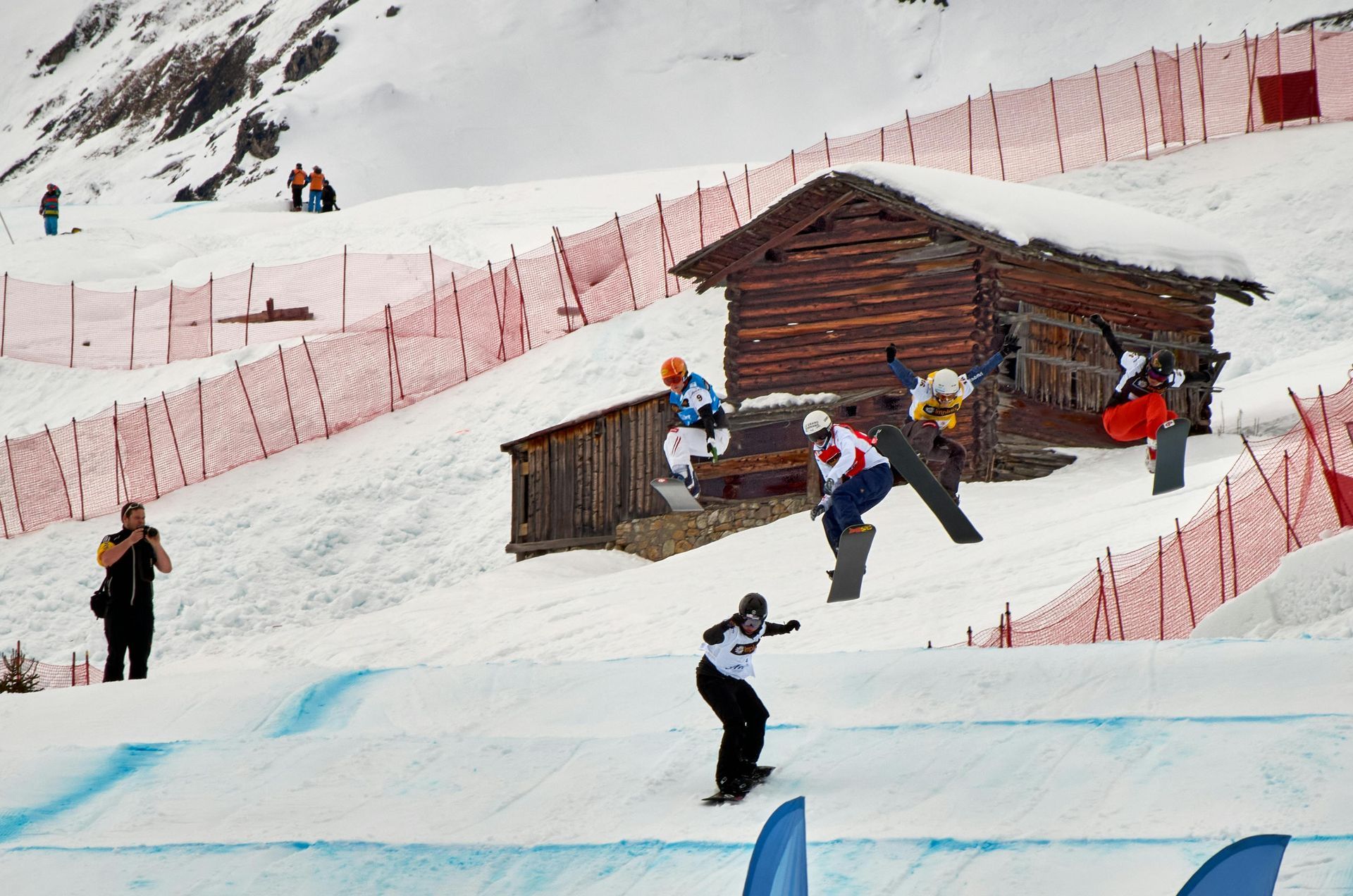 Snowboarders on a slope, jumping near a snow-covered wooden cabin. Red netting, snowy mountain backdrop.