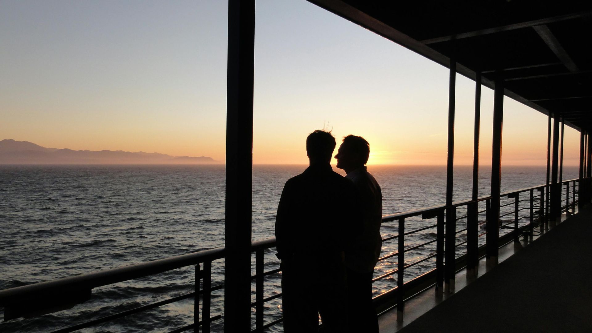 Two figures silhouetted on a ship deck, gazing at a sunset over the ocean.