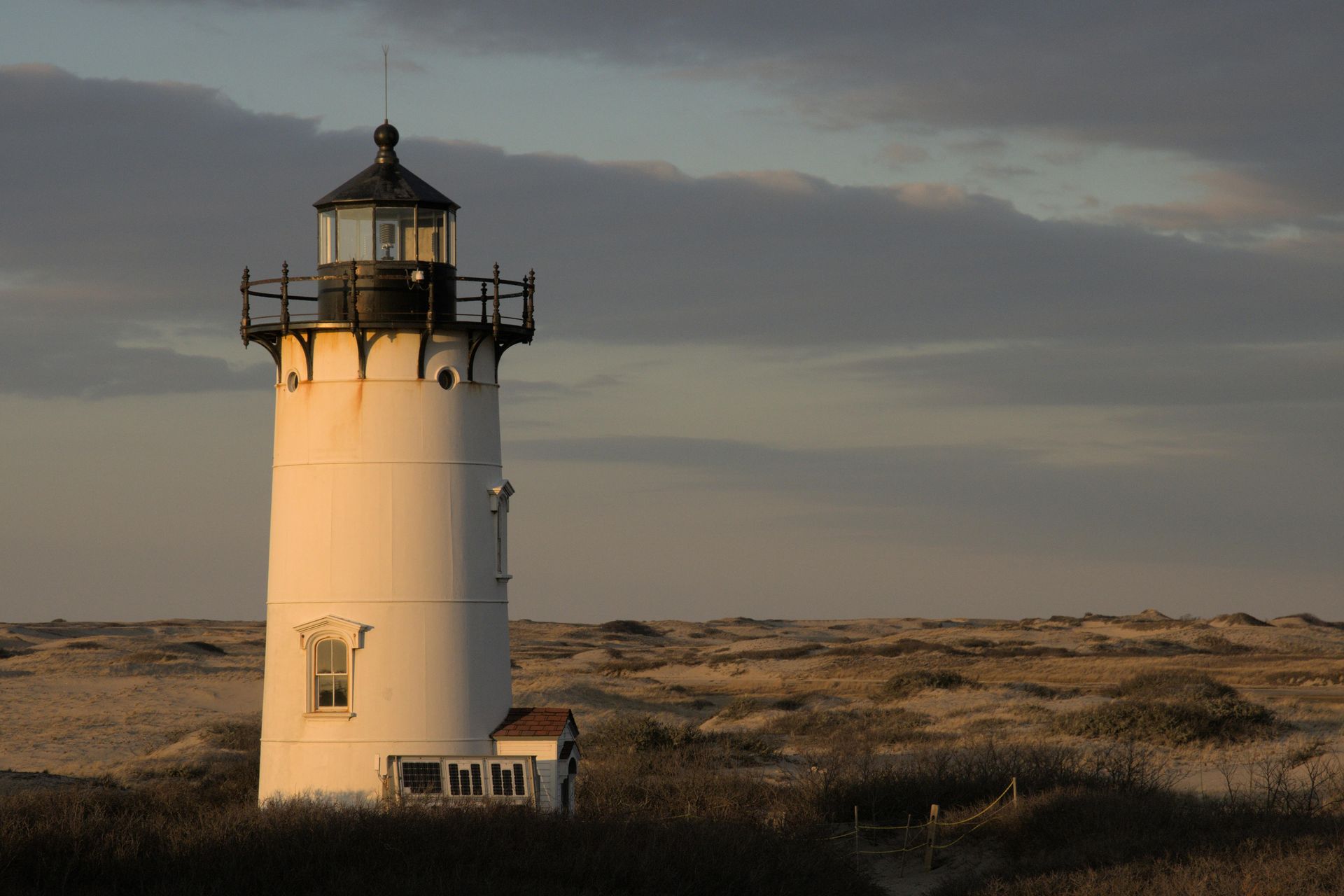 White lighthouse on a sandy dune under a cloudy sky, bathed in golden sunlight. Provincetown, USA: Coastal Cool