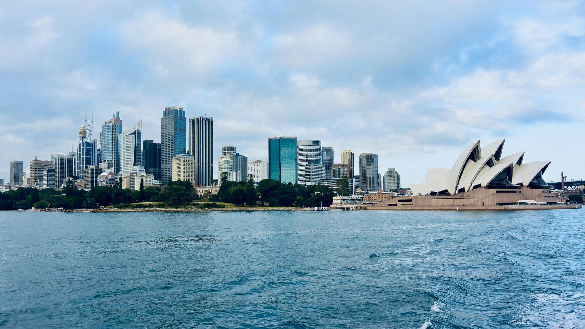 Sydney Opera House and city skyline viewed across water. Blue sky and choppy water.