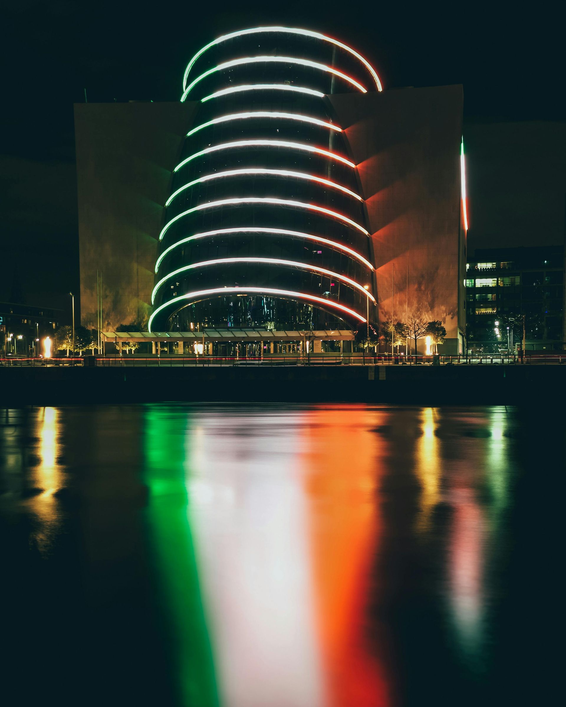 A tall, modern building with curved, glowing light strips, reflected in dark water with green, white, and orange hues.
