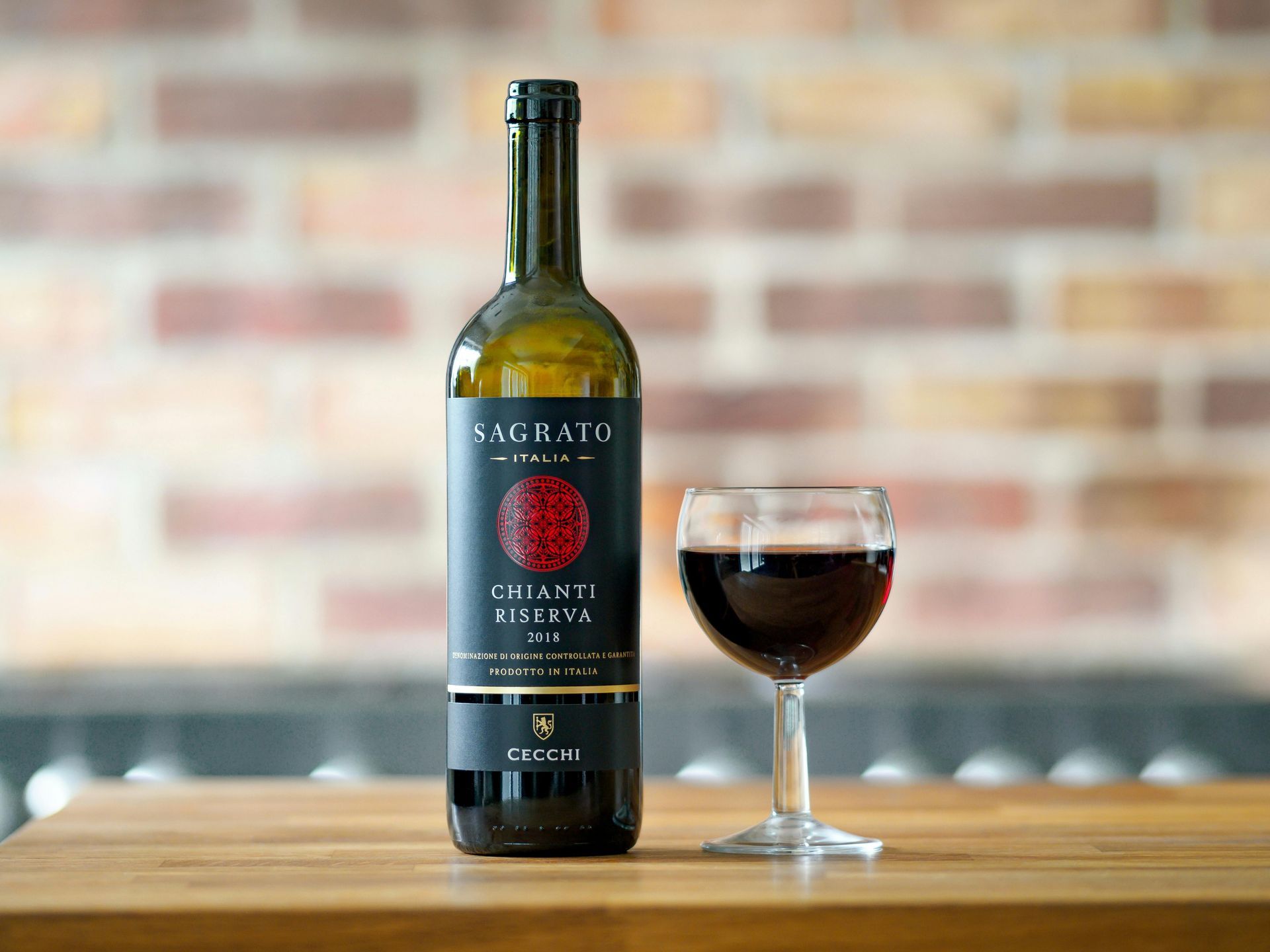 A bottle of Sagrato Chianti Riserva wine next to a glass of red wine on a wooden table against a brick wall.