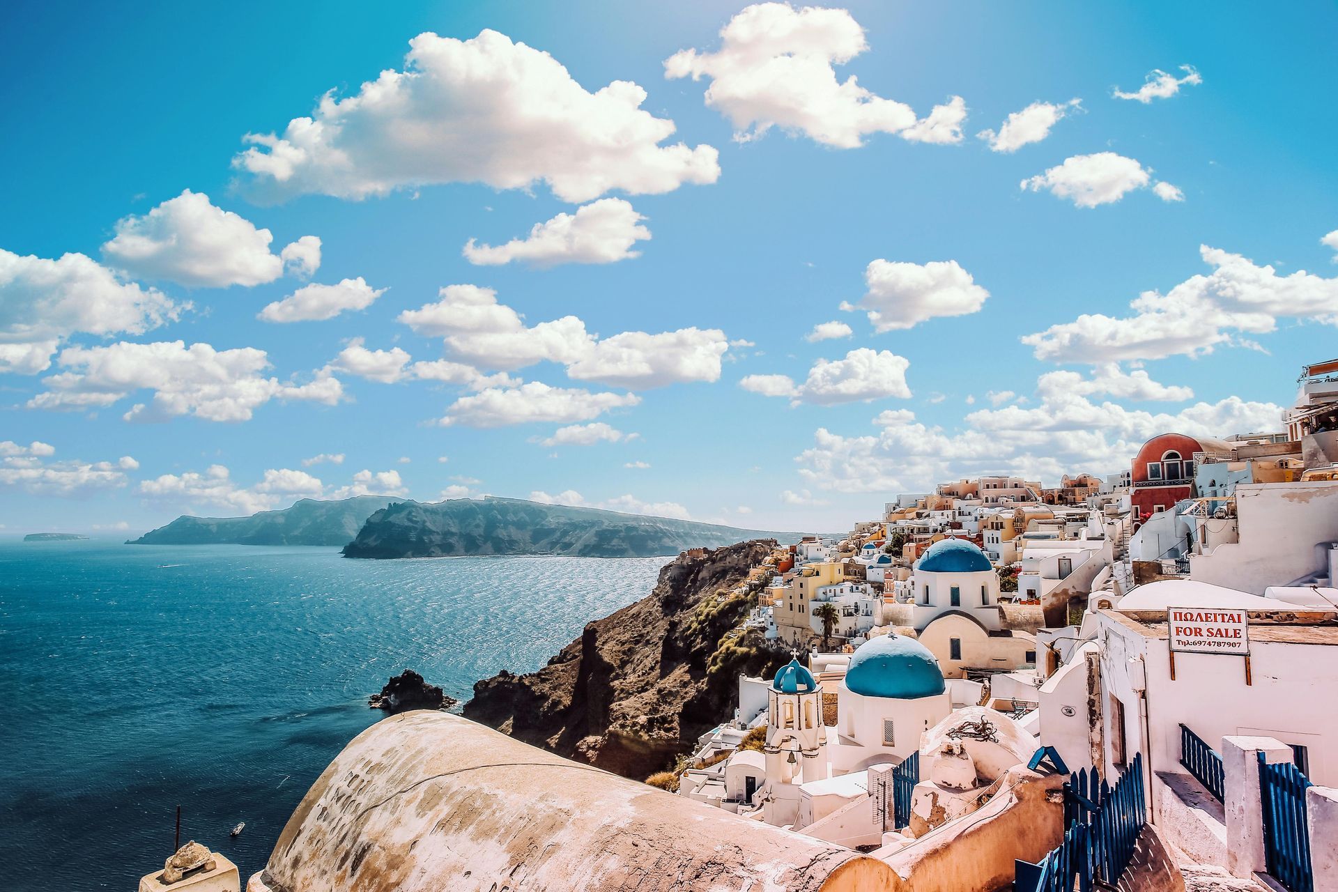 White buildings with blue domes on a cliff overlooking the Aegean Sea. Bright blue sky with fluffy clouds. Mykonos