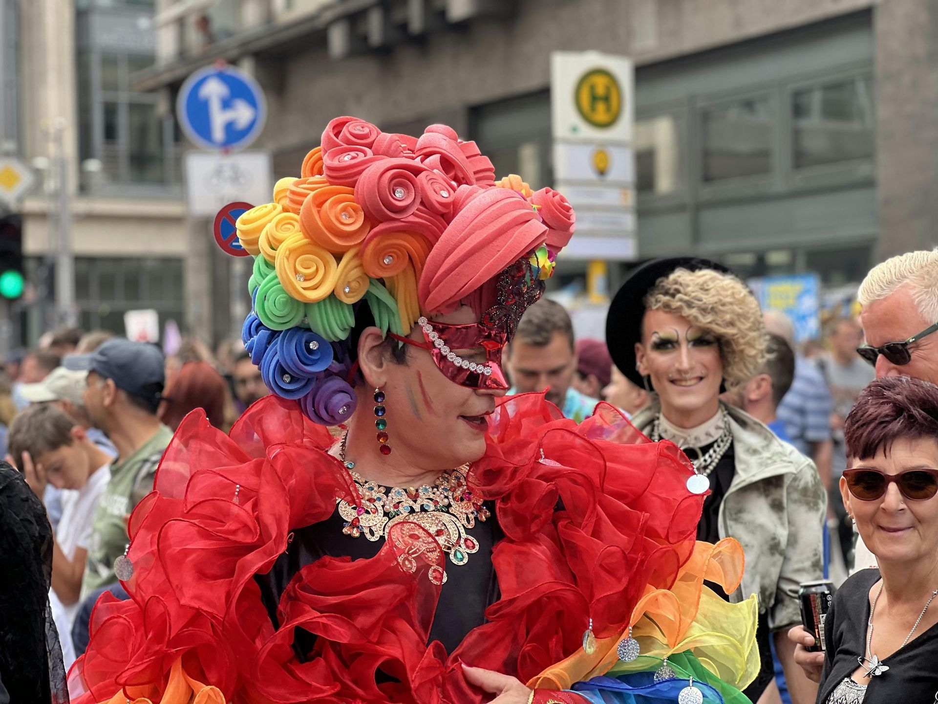 Person in elaborate rainbow-colored outfit at a Pride parade, with a red ruffled top and ornate headdress.