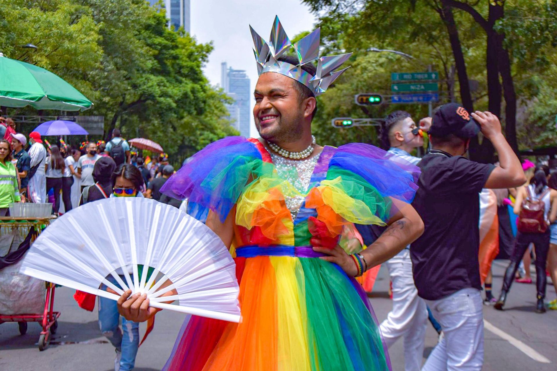 Person in rainbow dress and crown at Pride parade, holding fan.