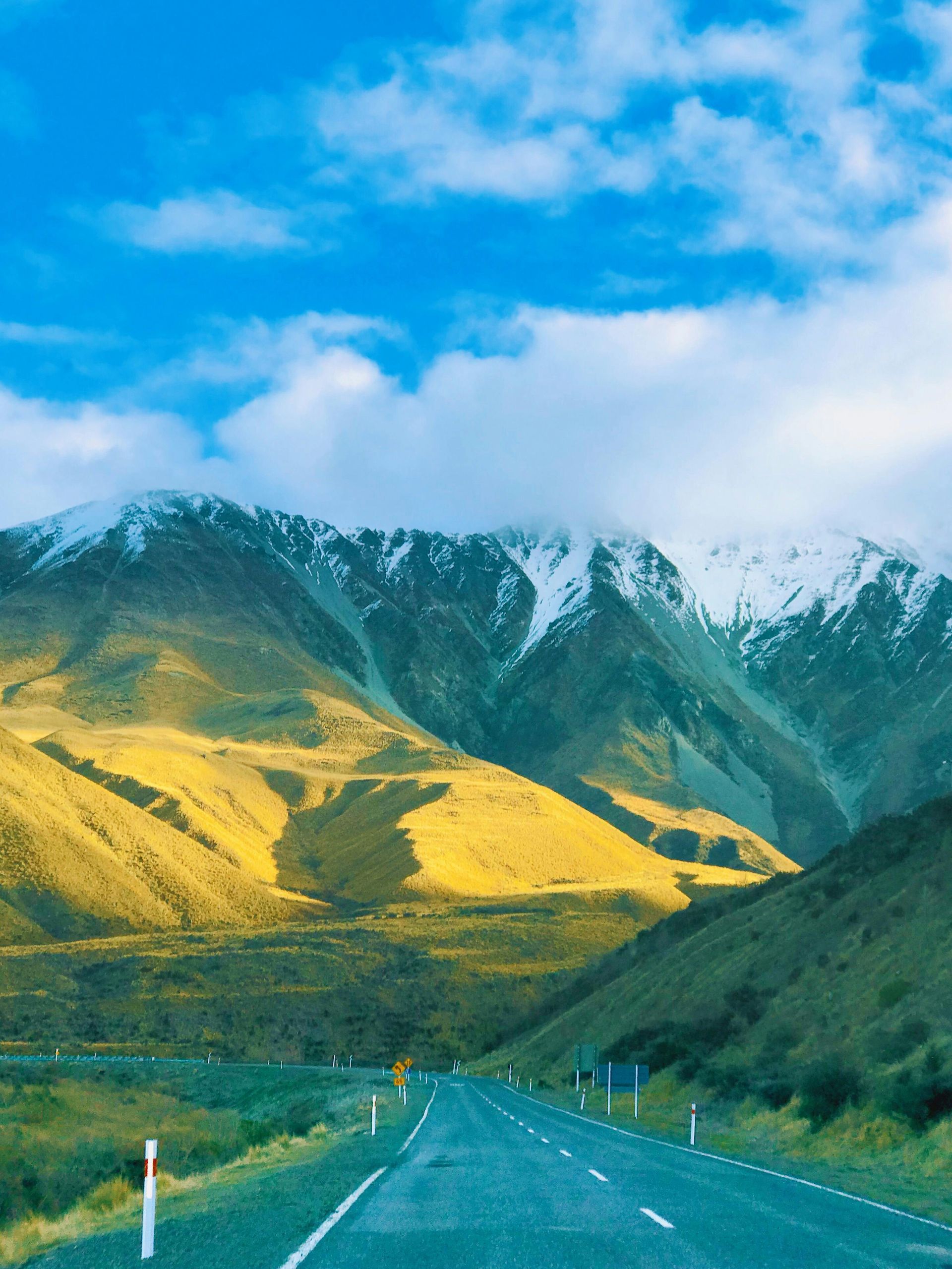 A road stretches toward sunlit, snow-capped mountains under a bright, cloudy blue sky.