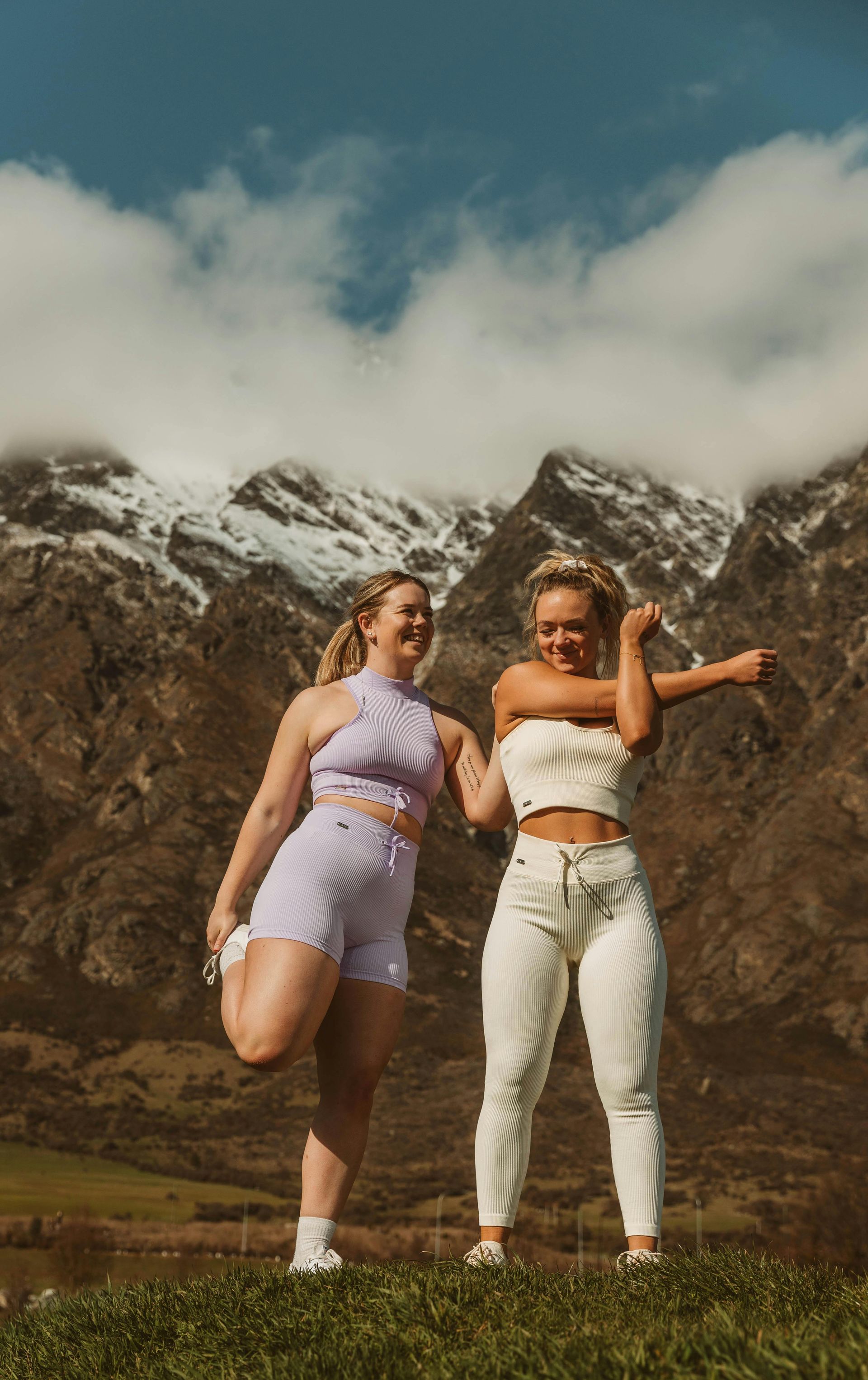Two people stretch in matching ribbed activewear against a backdrop of snow-capped mountains and a cloudy blue sky.