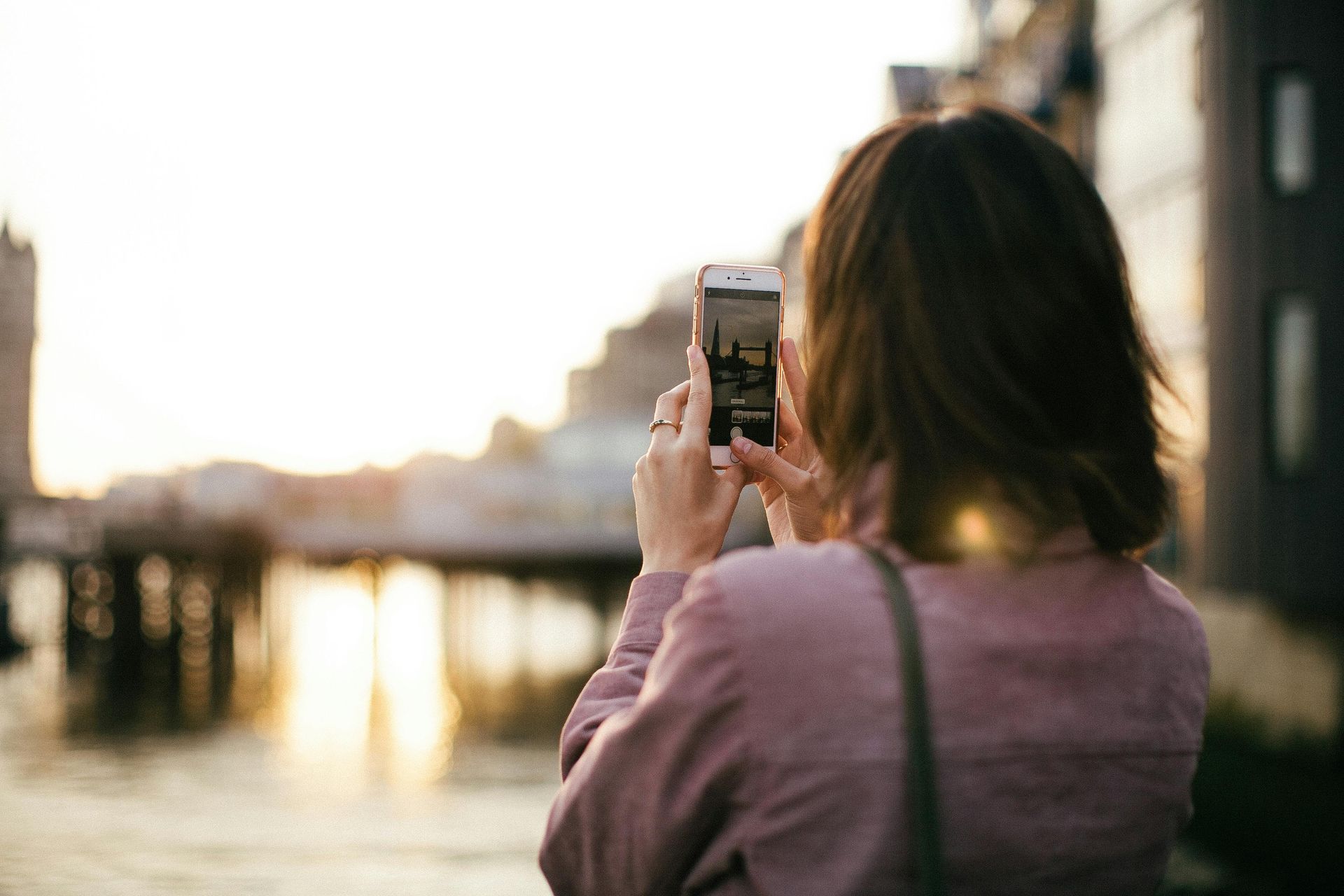 Woman taking photo with phone, backlit by a sunset over water.