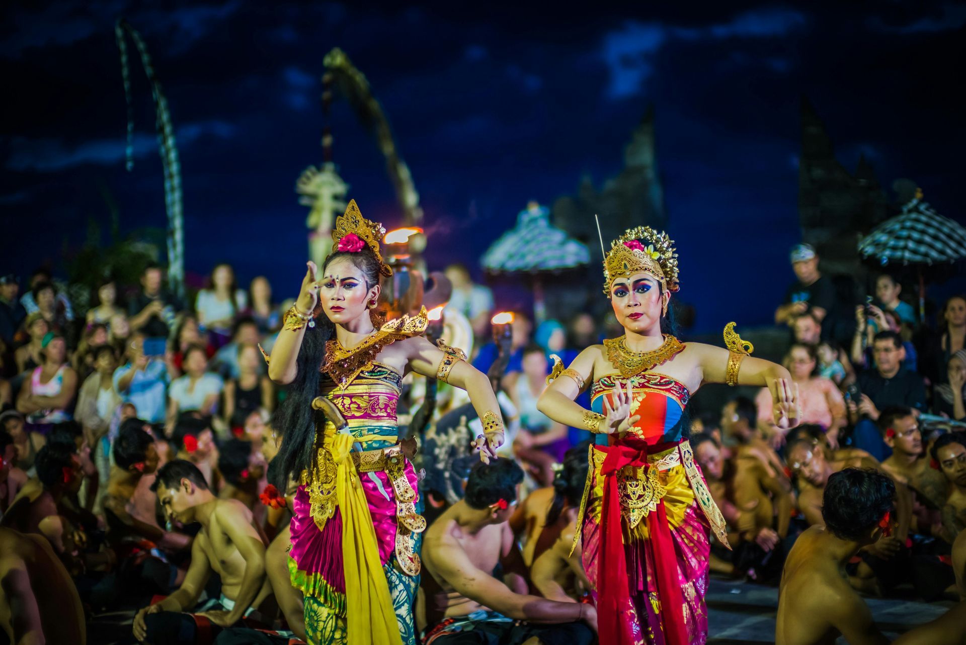 Balinese dancers performing in colorful costumes, outdoors at night. Audience watches.