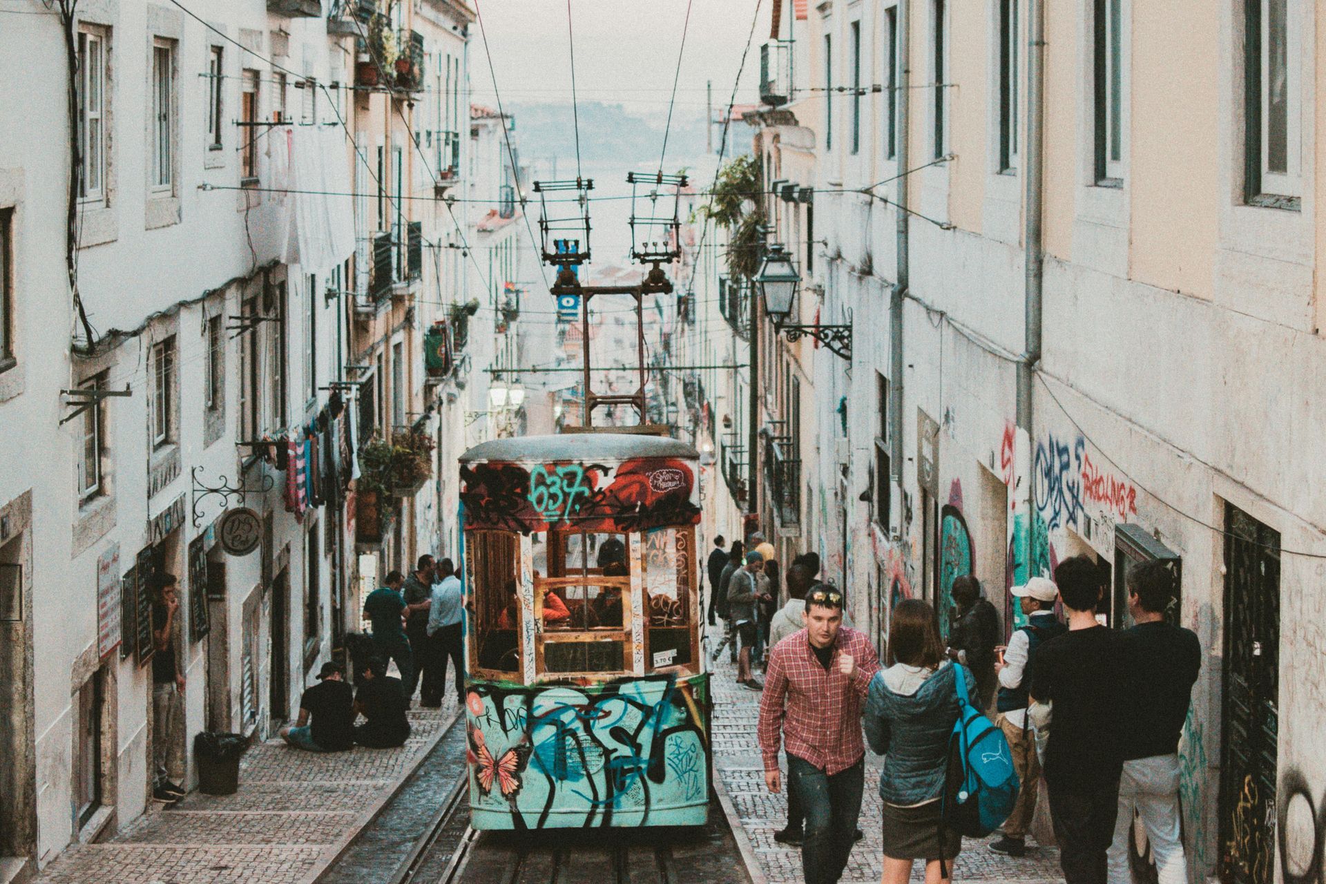 Cable car travels up a narrow, cobbled street lined with buildings. People stand nearby.