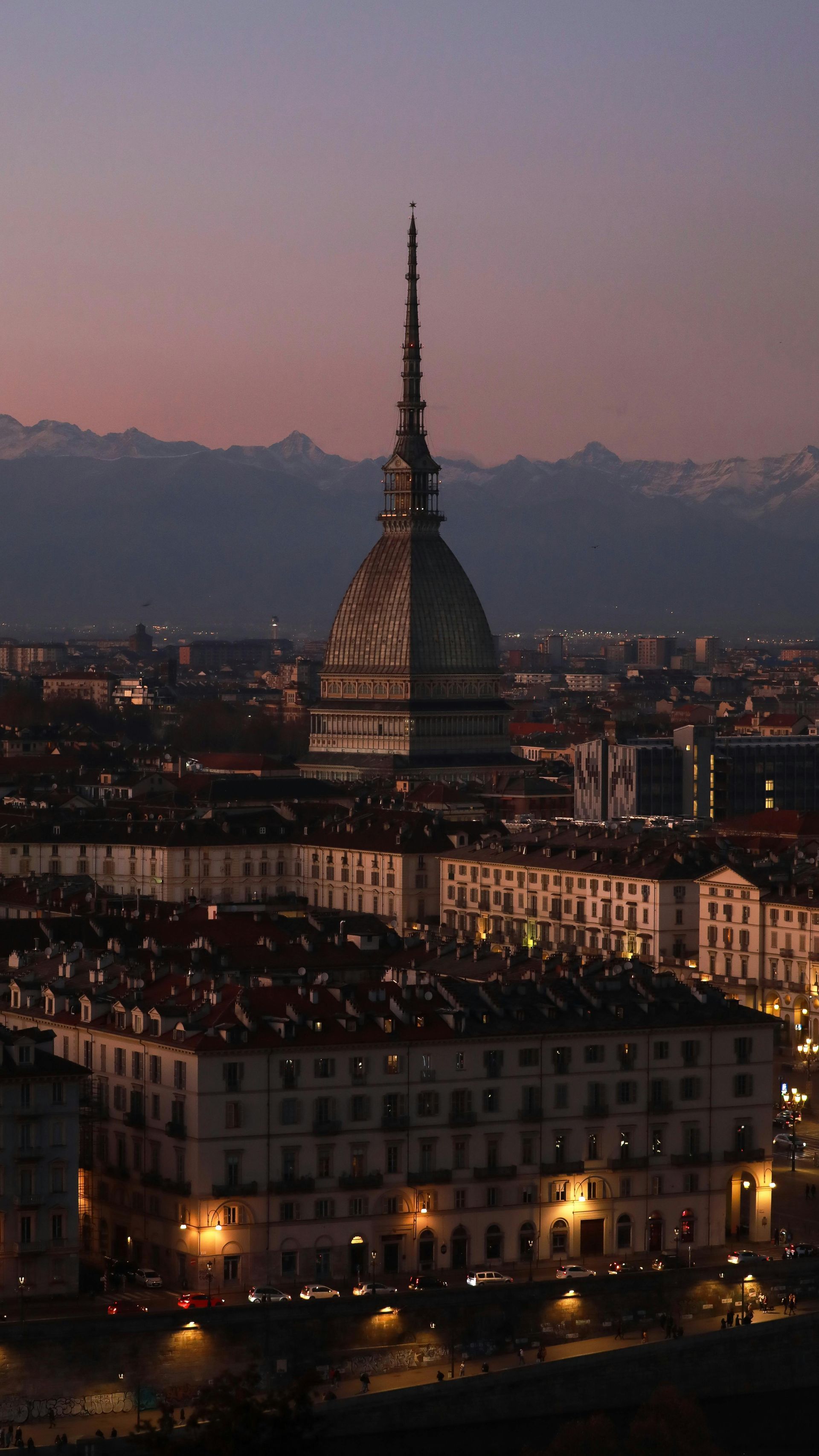 Mole Antonelliana, Turin, Italy, at dusk. The tall, spire-topped building towers over city rooftops with mountains in the background.