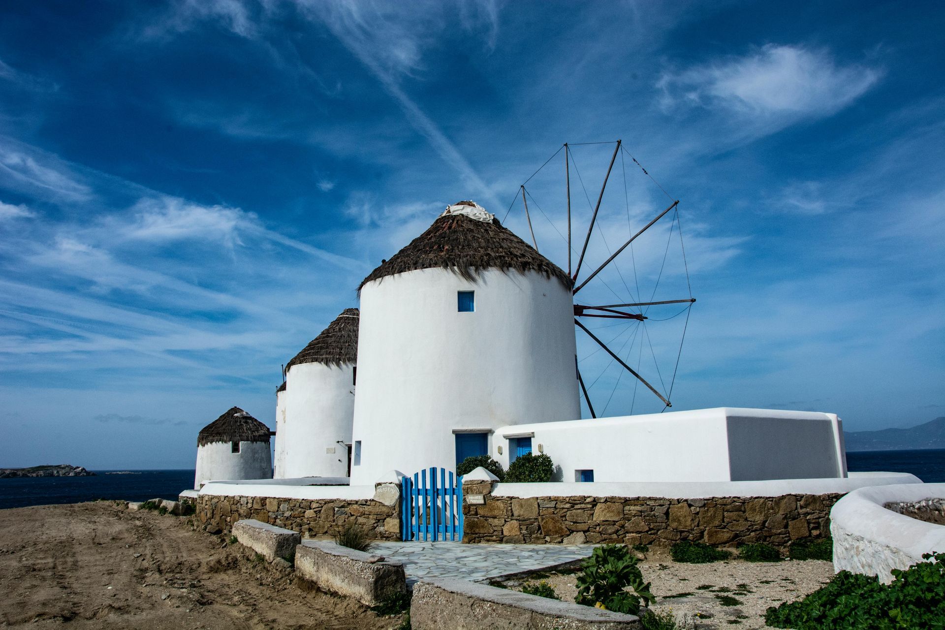 Three iconic white, stone windmills with thatched roofs overlooking the sea under a bright blue, cloudy sky on Mykonos.
