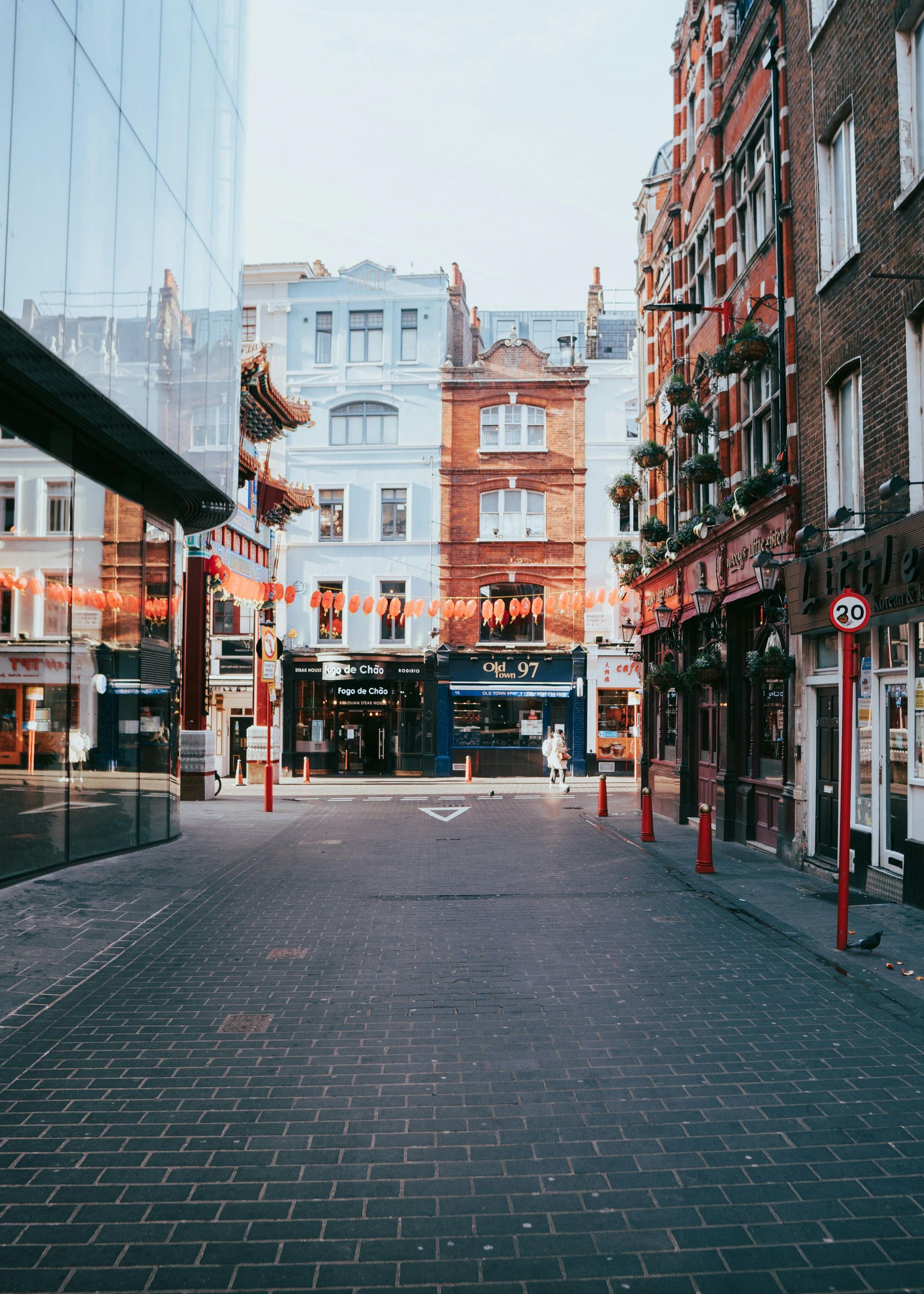 Narrow street lined with brick buildings, shops, and red poles. Blue sky overhead. Soho – London