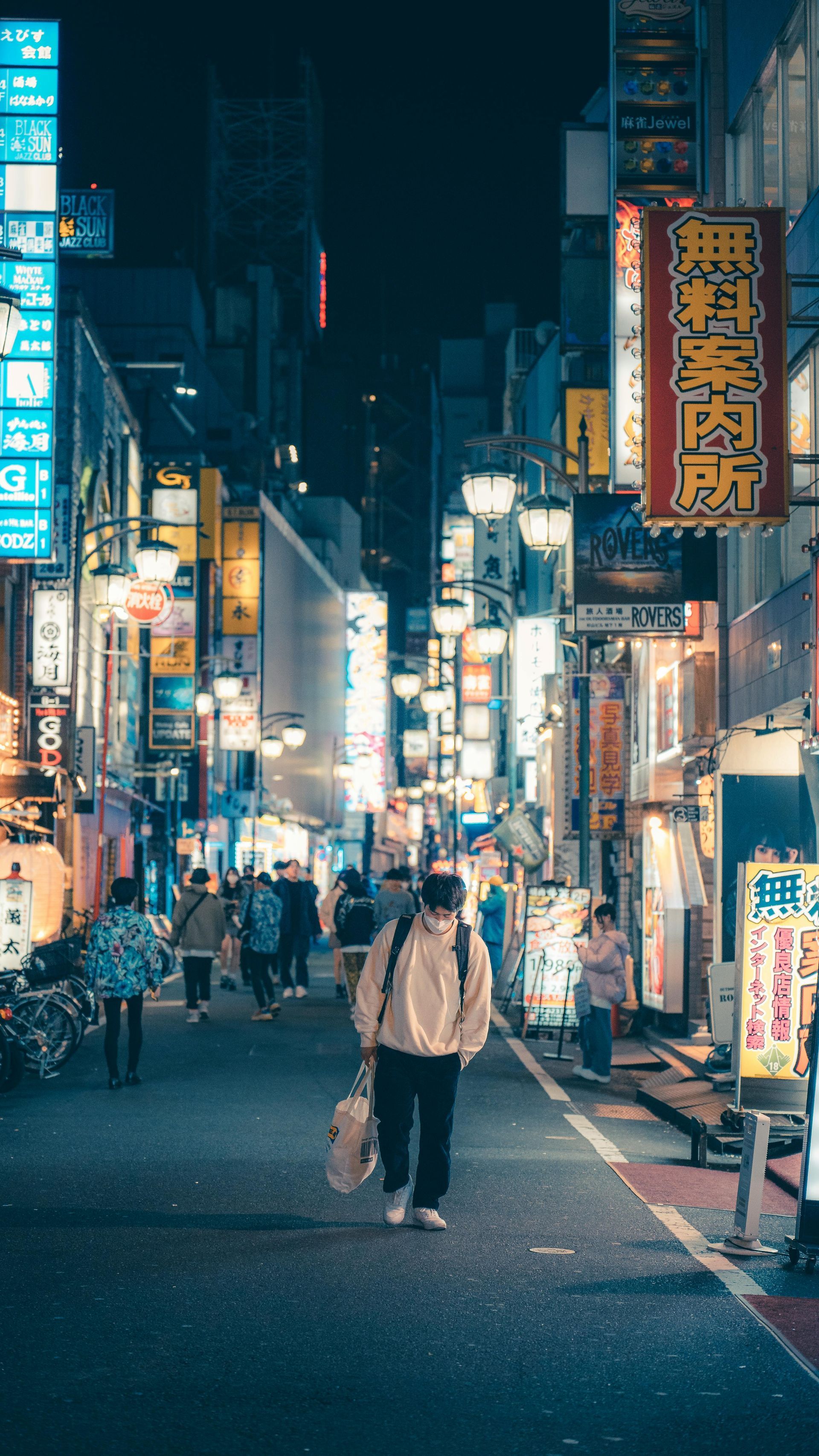 Person walks down a vibrant, neon-lit street in Japan at night. Many signs and buildings. Shinjuku Ni-chōme – Tokyo